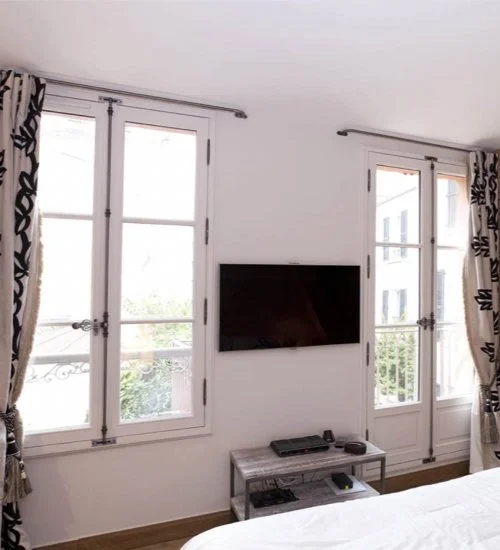 Une pièce avec deux fenêtres doubles et un téléviseur mural, avec un meuble en dessous tenant une console et une télécommande, et des rideaux à motifs noirs et blancs.