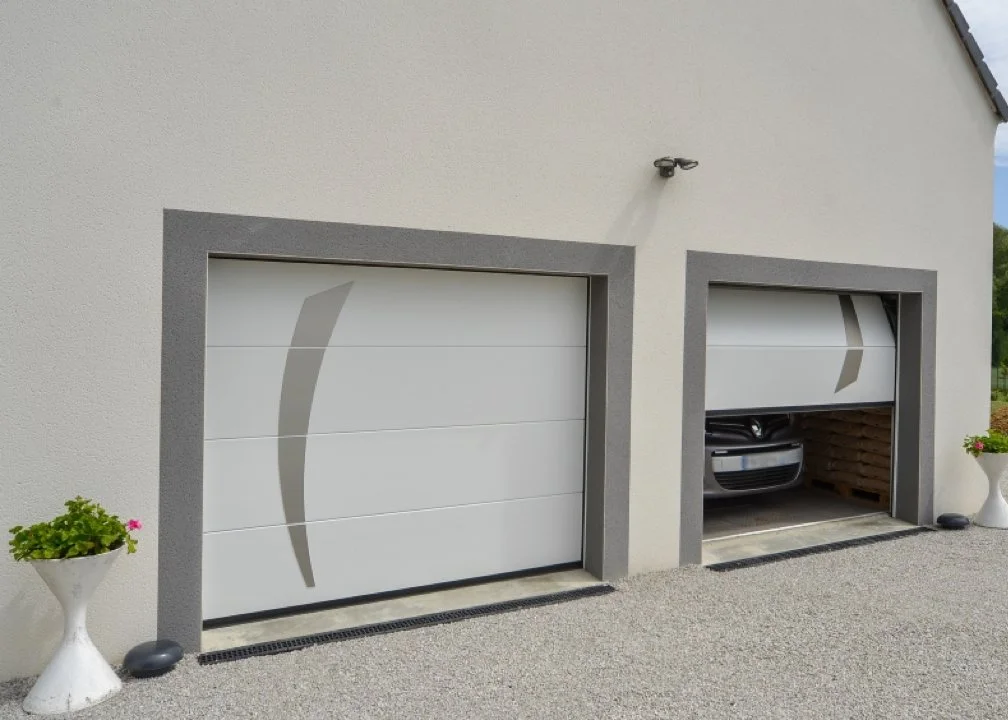 Deux garages avec portes sectionnelles, l'un fermé et l'autre partiellement ouvert, avec voiture garée à l'intérieur, devant un mur en béton et entouré de pots de fleurs.