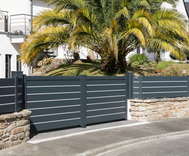 Une clôture en métal gris foncé devant un jardin avec un palmier et d'autres plantes, bâtiment blanc en arrière-plan.