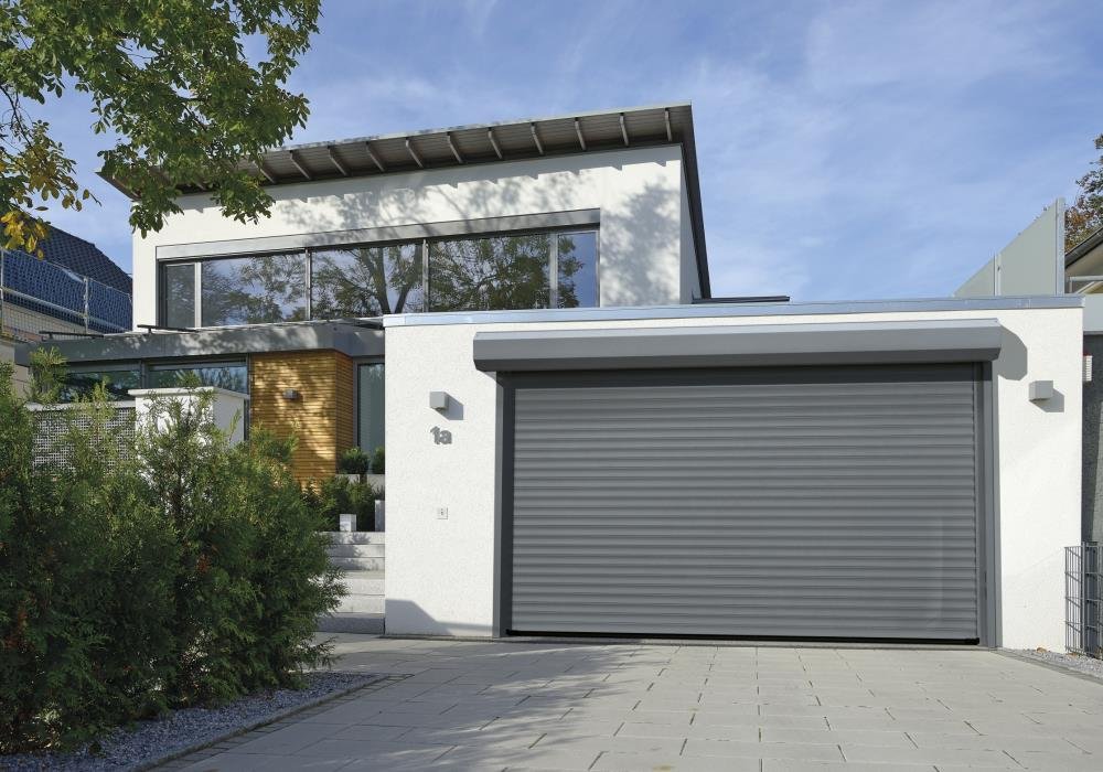 Une maison moderne avec un garage à volet roulant gris et une entrée principale avec des marches en béton, entourée de végétation et sous un ciel bleu.