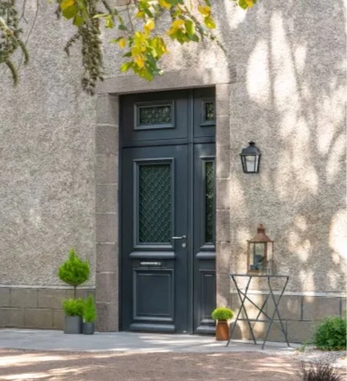 Porte noire élégante avec fenêtres en verre décoratif, entourée d'un mur en pierre et décorée avec des pots de plantes vertes et une lanterne d'extérieur.