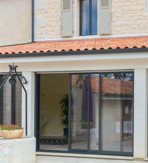 Façade de maison avec fenêtres à volets, porte-fenêtre coulissante en verre, toit en tuiles orangées, mur en pierre beige, et un lanterne de jardin.