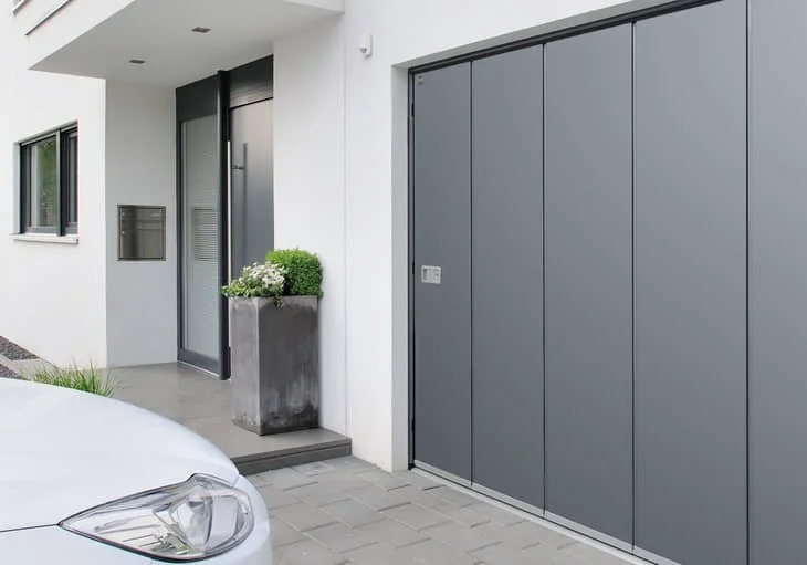 Facade moderne d'une maison avec porte d'entrée, fenêtres, un grand garage gris et un pot de fleurs avec des plantes vertes.
