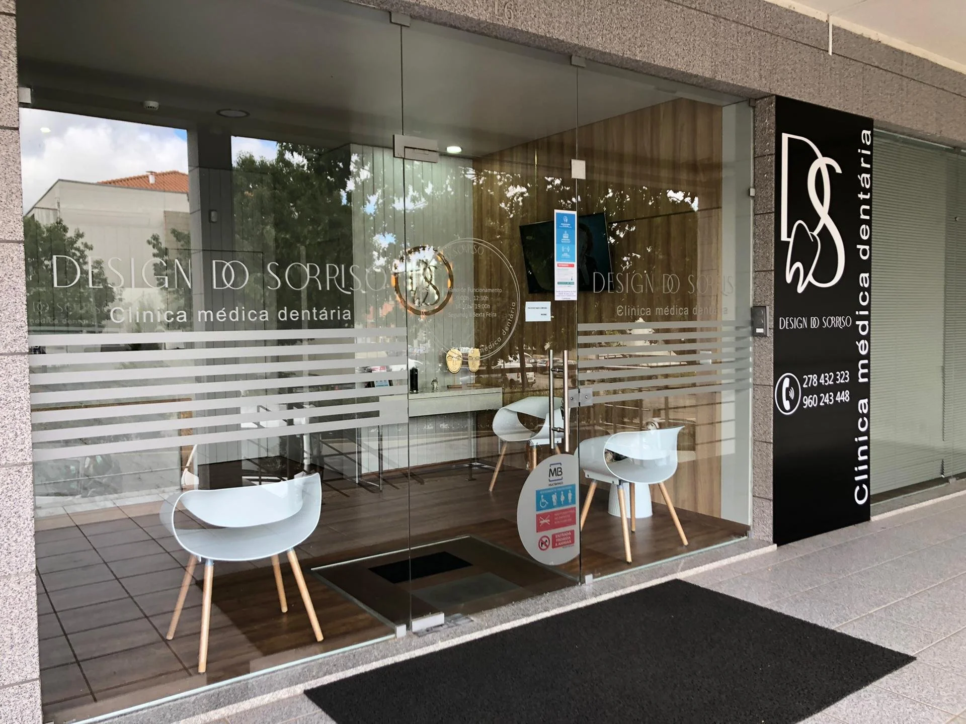 Fachada de uma clínica odontológica chamada 'Design do Sorriso', com vidro transparente, duas cadeiras brancas na entrada, logo da clínica com desenho de um sorriso, e informações de contato na lateral preta.