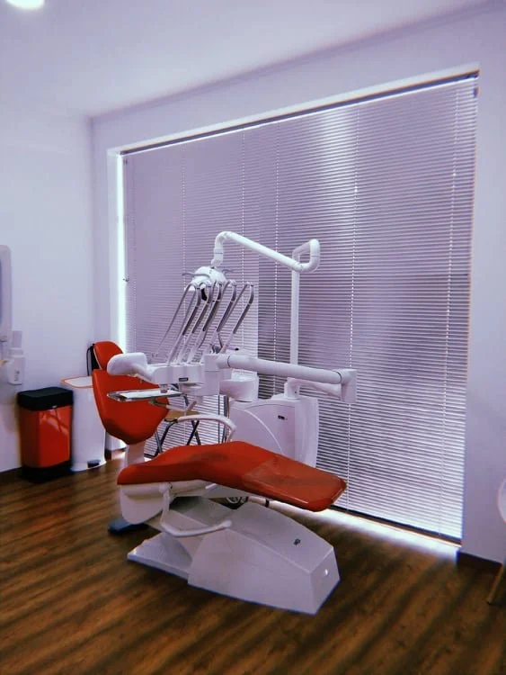 Cadeira odontológica vermelha em consultório com janela com persianas fechadas.