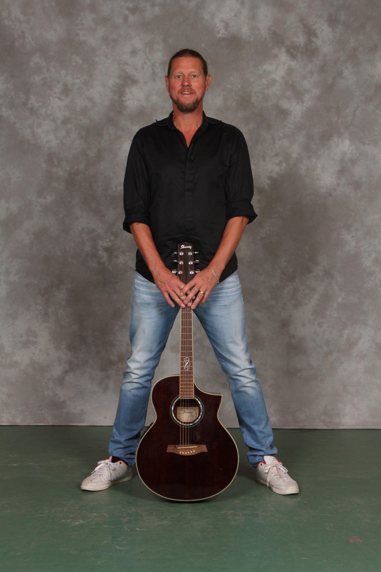 Per Hilmersson står framför en grå bakgrund, håller en akustisk gitarr framför honom, ser glad ut, bär svart skjorta och jeans.