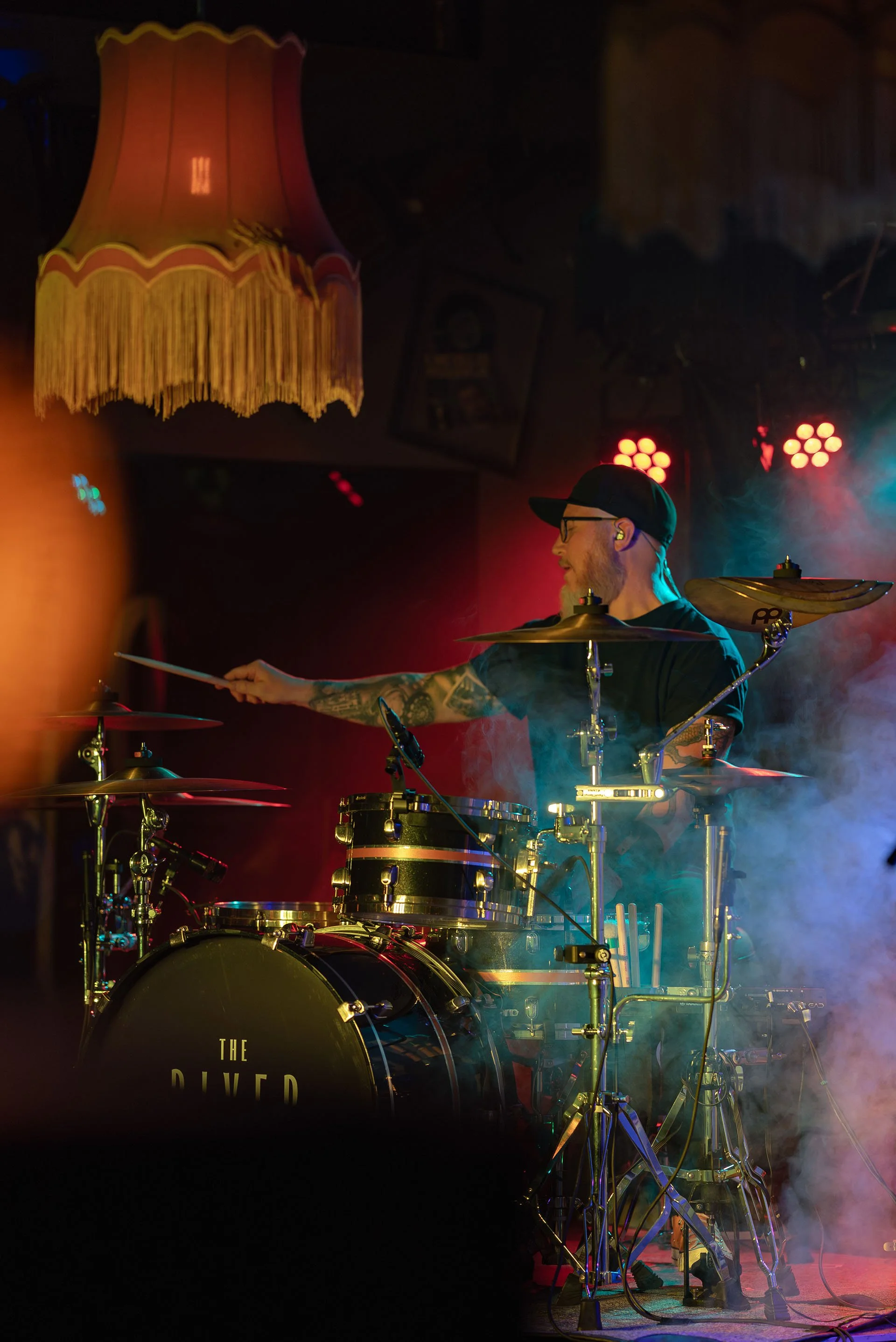 En man som spelar trummor på en scen, med färgglad scenbelysning och rök, samt en vintage taklampa hängande från taket.
