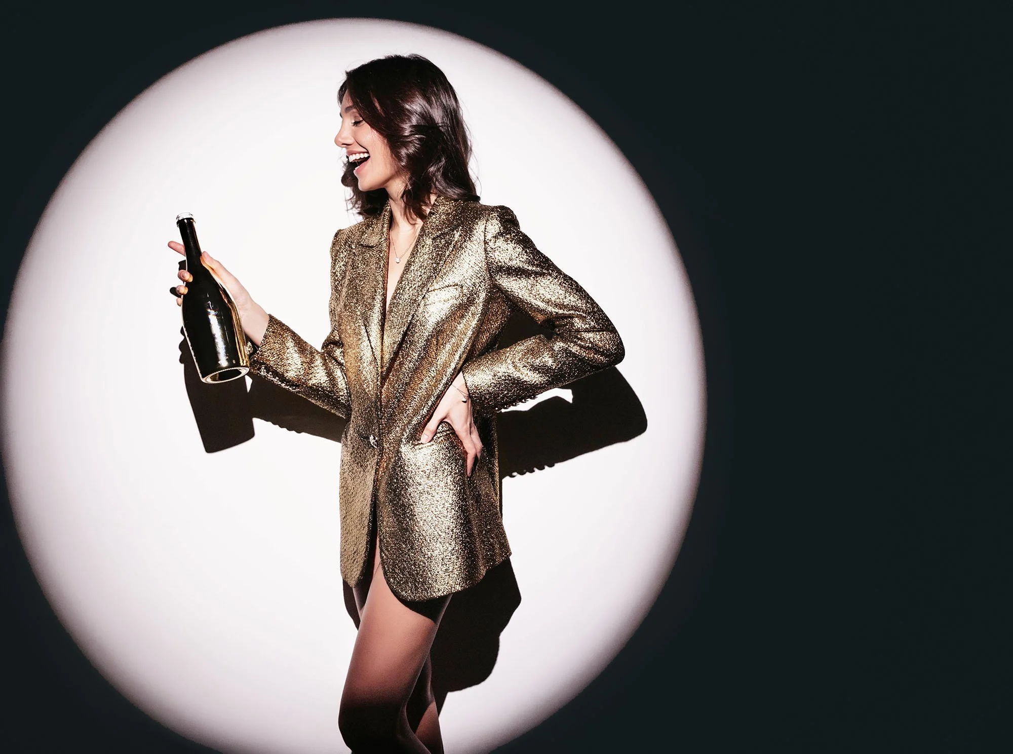 En kvinna i en guldglittrande kavaj håller en flaska i handen och skrattar.