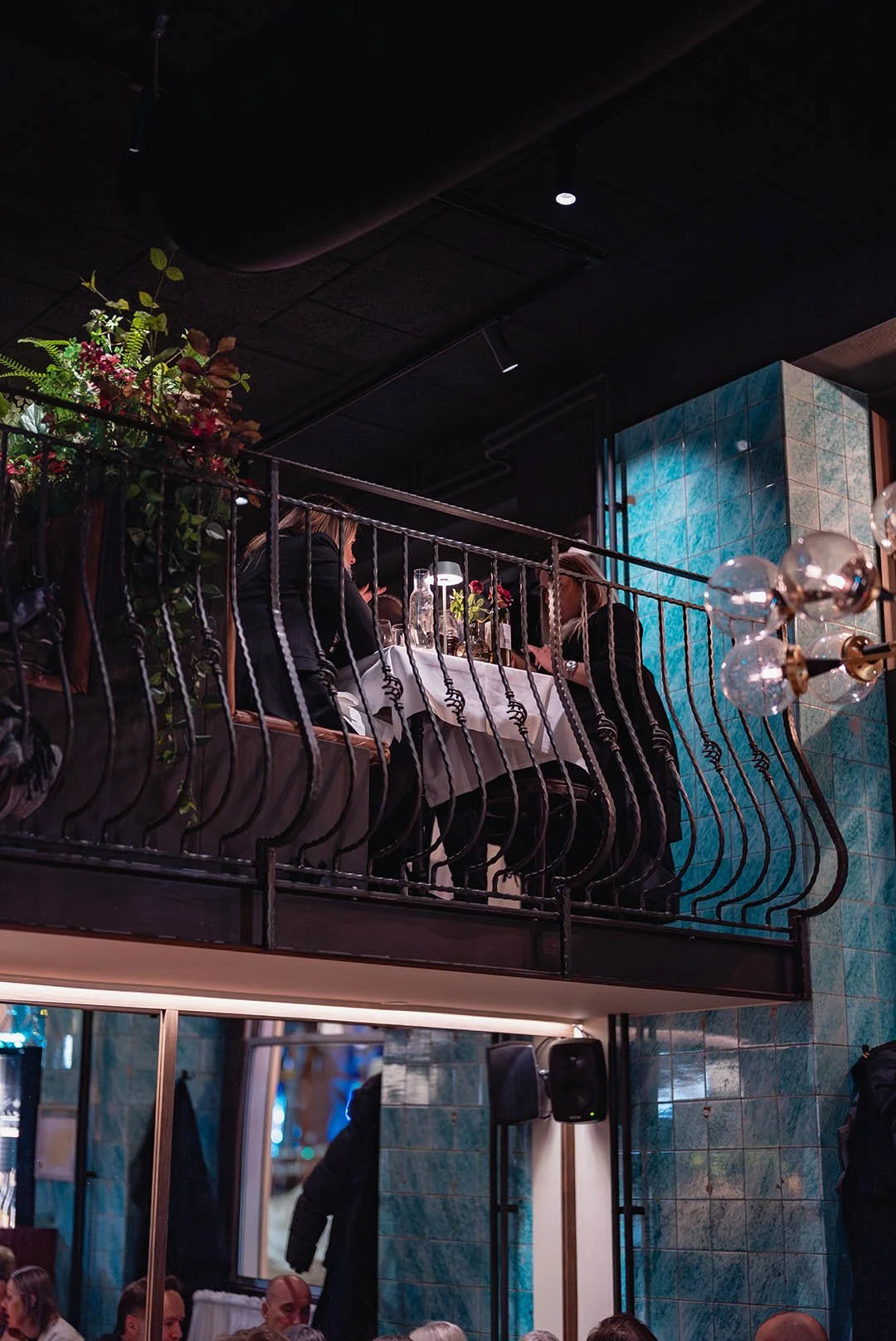 A restaurant scene with people dining at a table on an elevated balcony. The picture is taken from below, showing a metal railing and greenery decoration on the balcony. The interior features dark ceilings, teal tiled walls, and modern lighting.