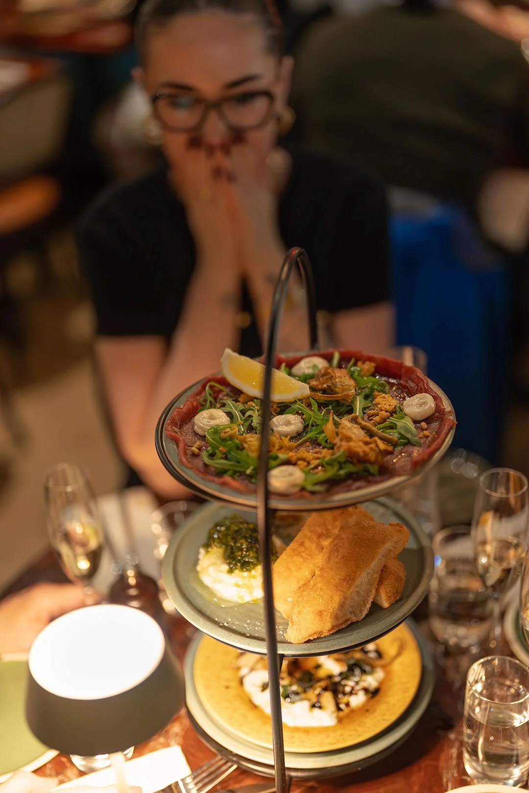 A woman with glasses and earrings gazes at a three-tiered serving stand filled with various dishes, with her hands clasped near her face.