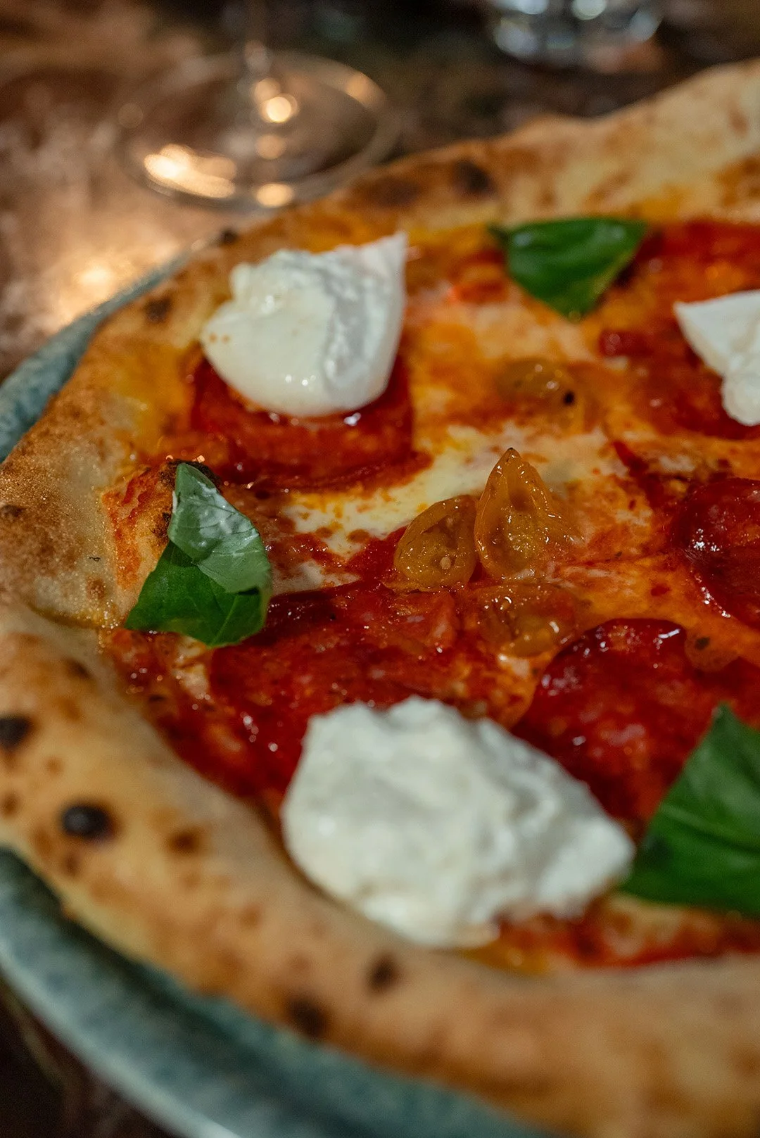 Close-up of a pizza with pepperoni, cherry tomatoes, basil leaves, and dollops of ricotta cheese on a pizza crust.