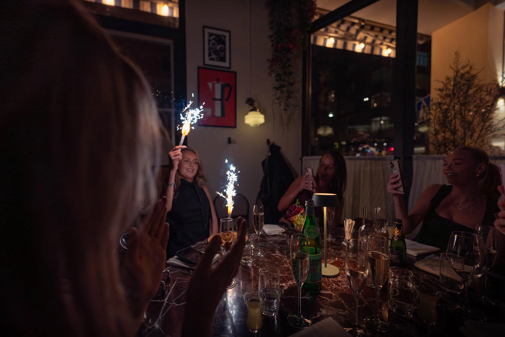 People celebrating at a dinner table with sparklers, taking photos, and smiling in a dimly lit restaurant or party room.
