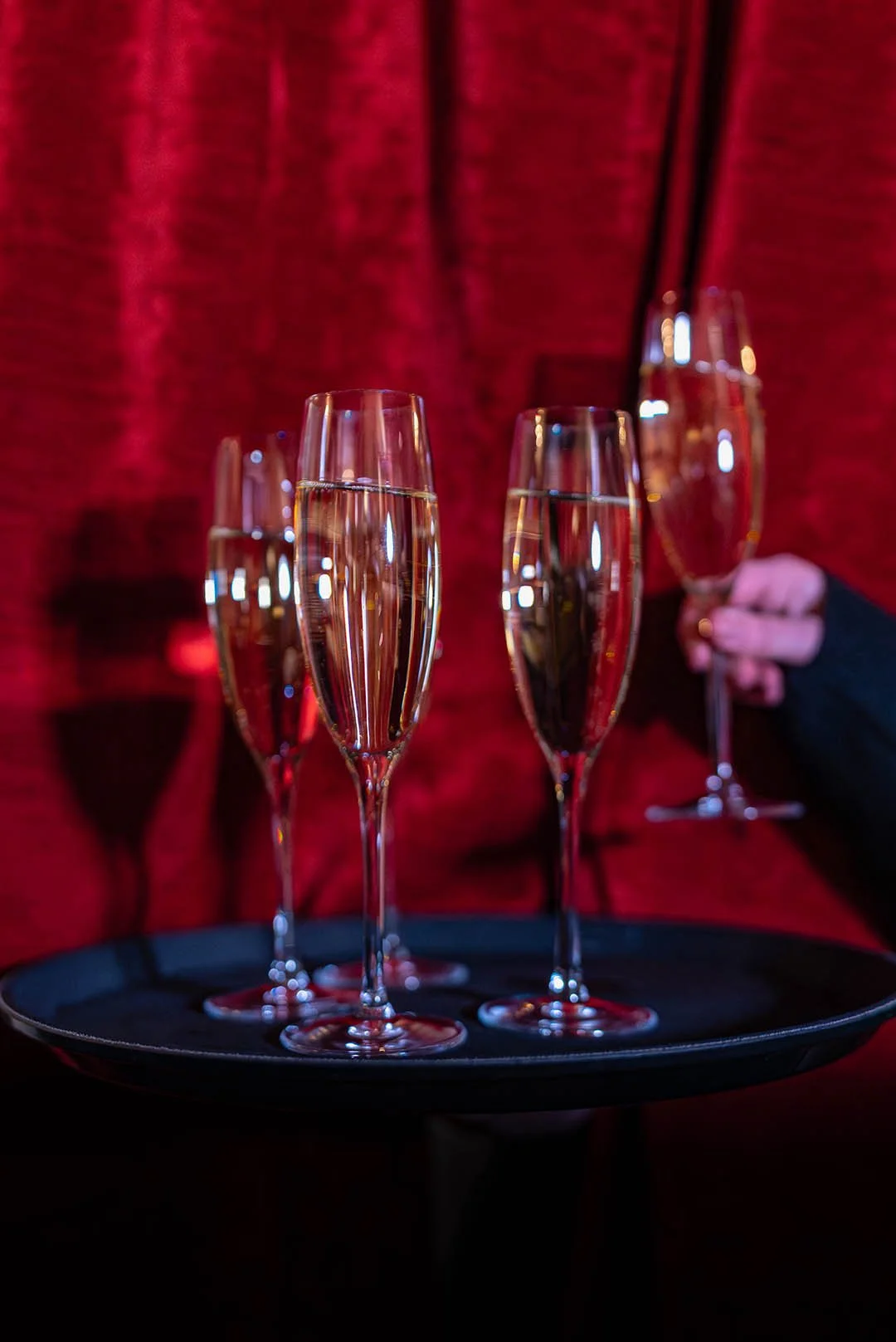 Four glasses of champagne on a tray, with a person holding one glass in the background against a red curtain.