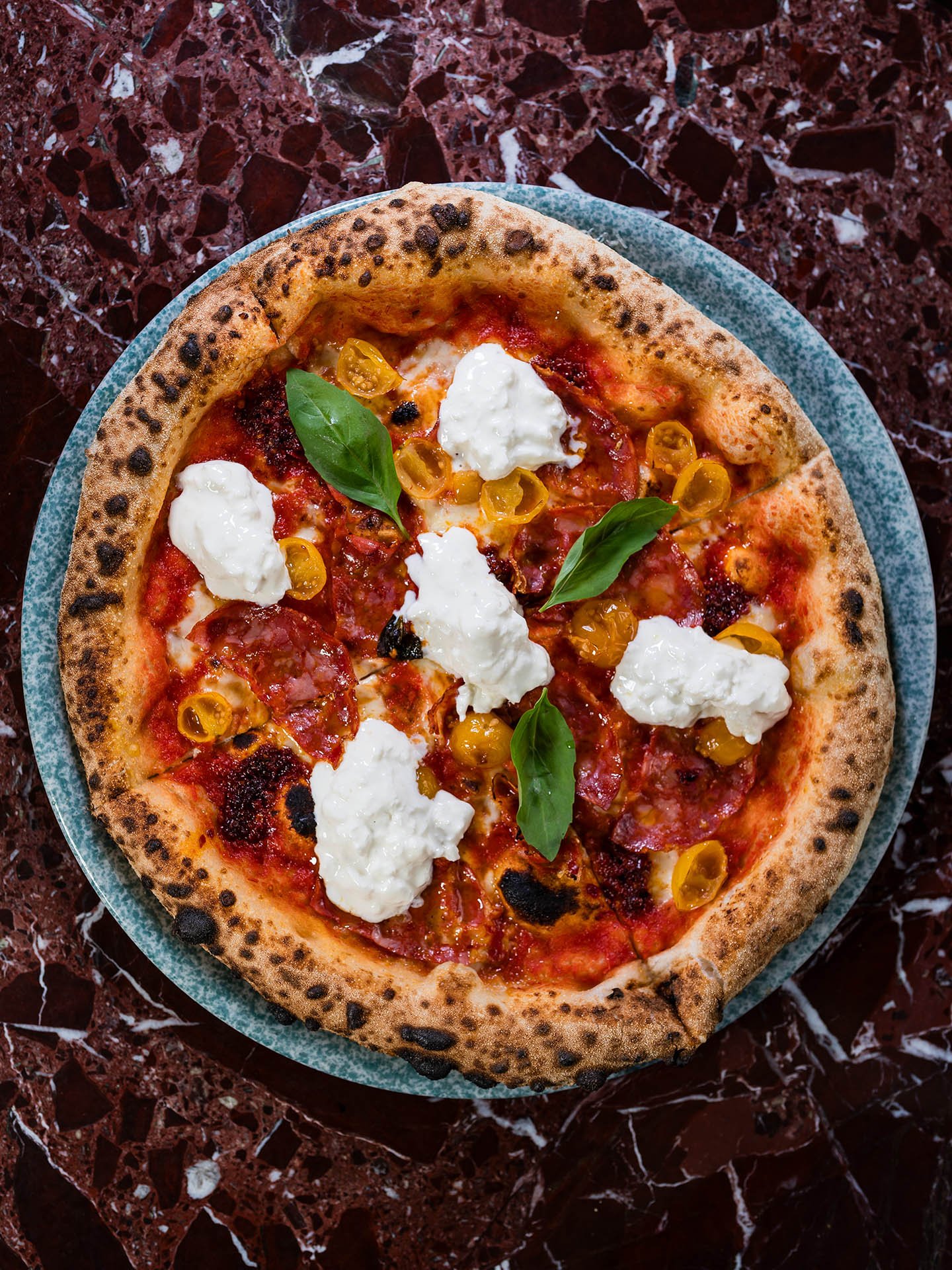 Tom pizza med mozzarella, skivor av chorizo och färska basilkblad på en grön assiett på en mörk marmorbord.