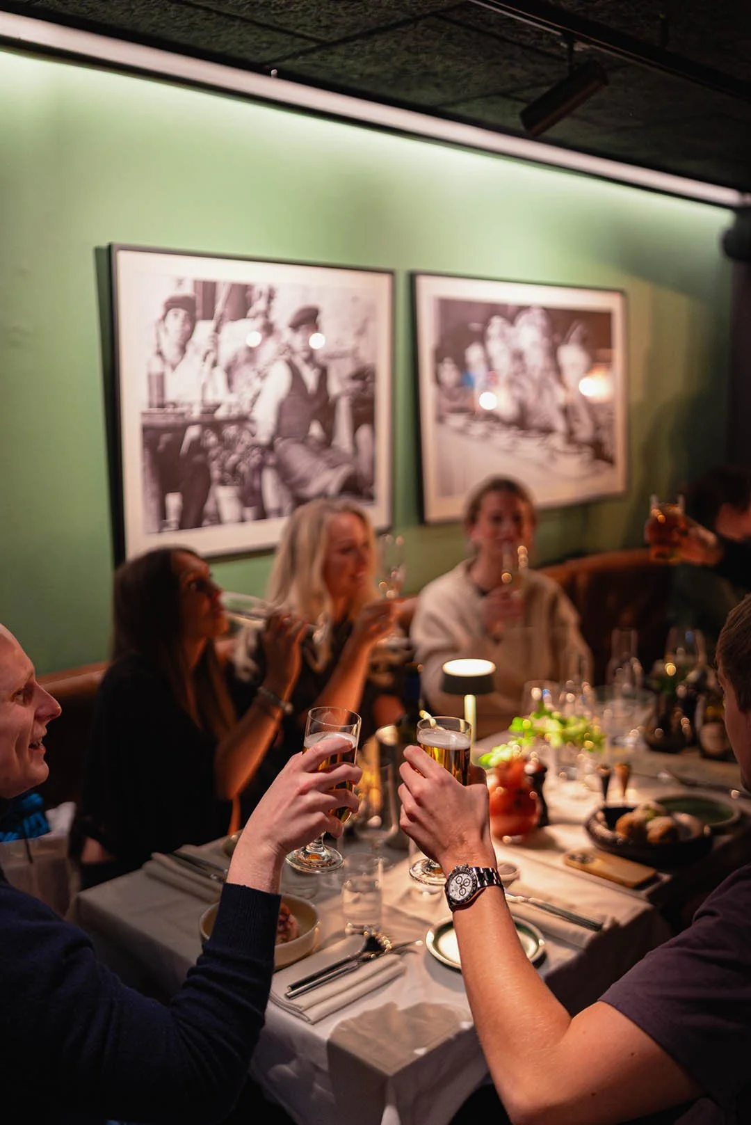 People celebrating at a dinner party with wine glasses in a restaurant decorated with black and white photographs on a green wall.