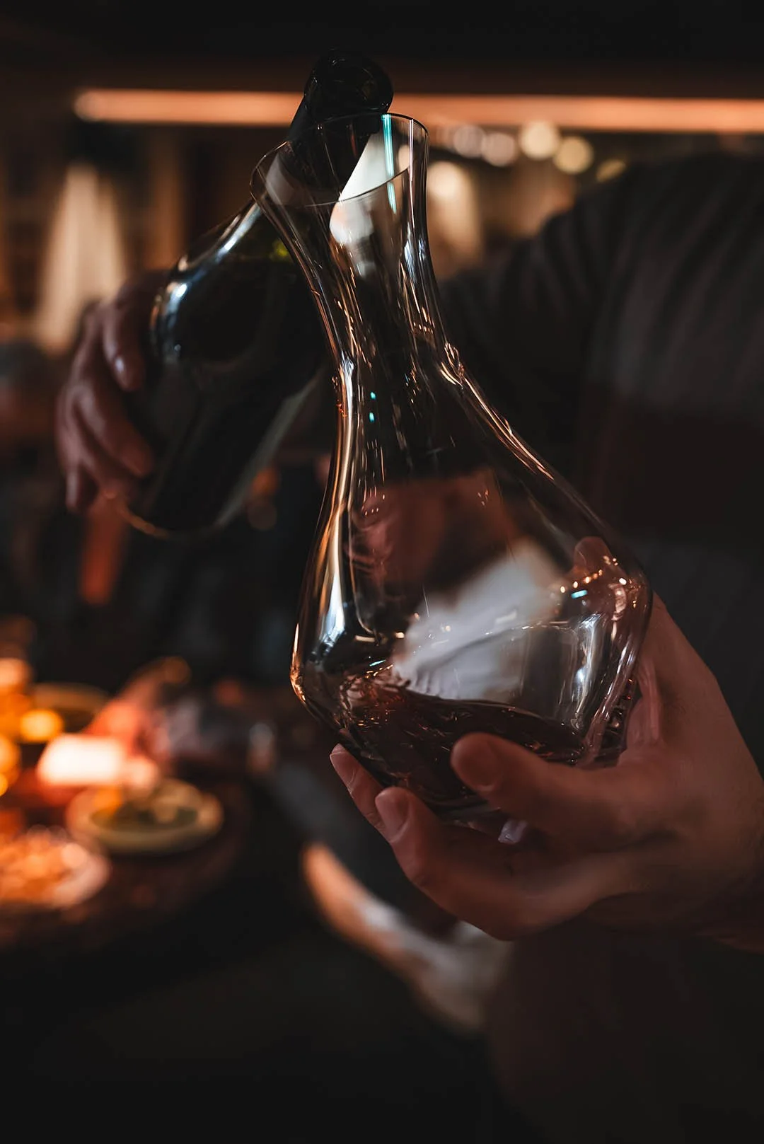 Person pouring red wine from a decanter into a glass in a dimly lit setting.