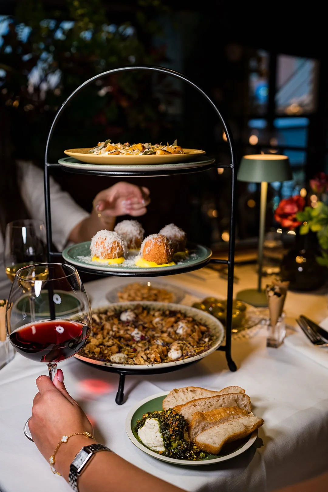 En trevånings serveringsställ med mat och en person som håller ett glas rött vin i en elegant middagsmiljö.