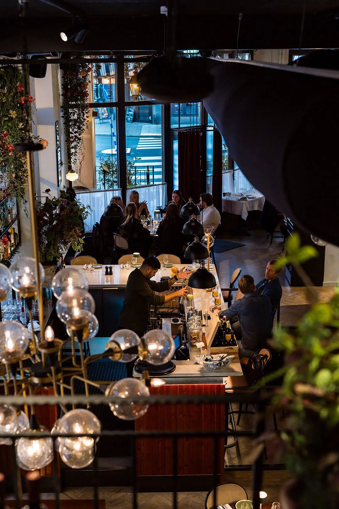 Interiör av en modern restaurang med flera bord, personal som serverar drinkar och gäster som äter och pratar, stora fönster med utsikt över trottoaren utomhus.