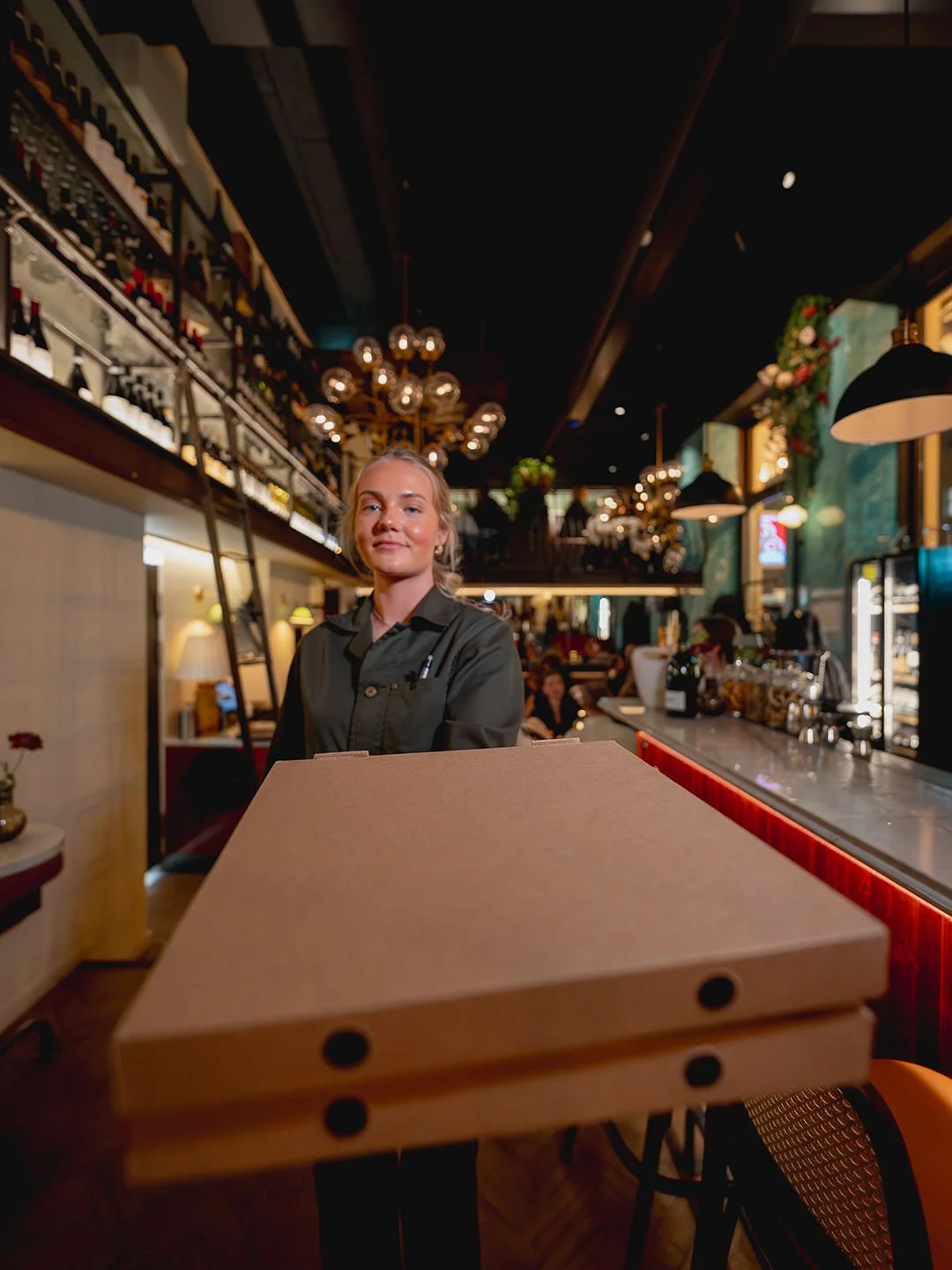 Young woman restaurant server with smiling face standing behind a pizza box at a bar or restaurant interior.