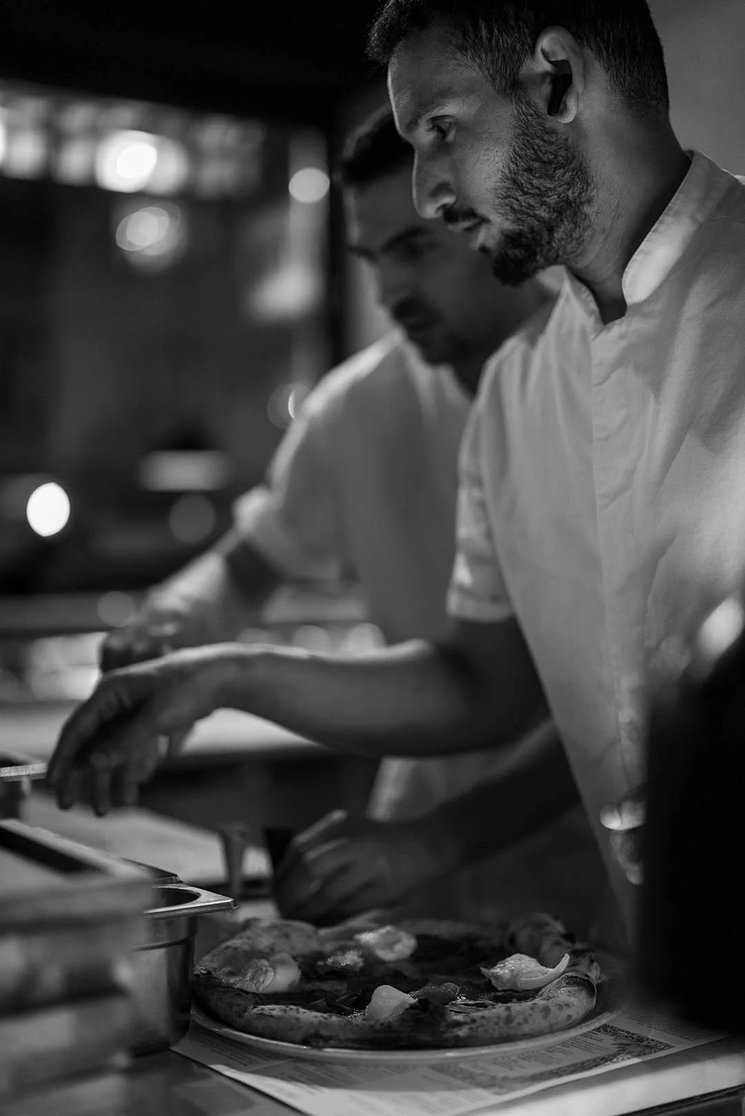 Two chefs in white uniforms preparing a pizza in a professional kitchen, focusing on adding toppings.