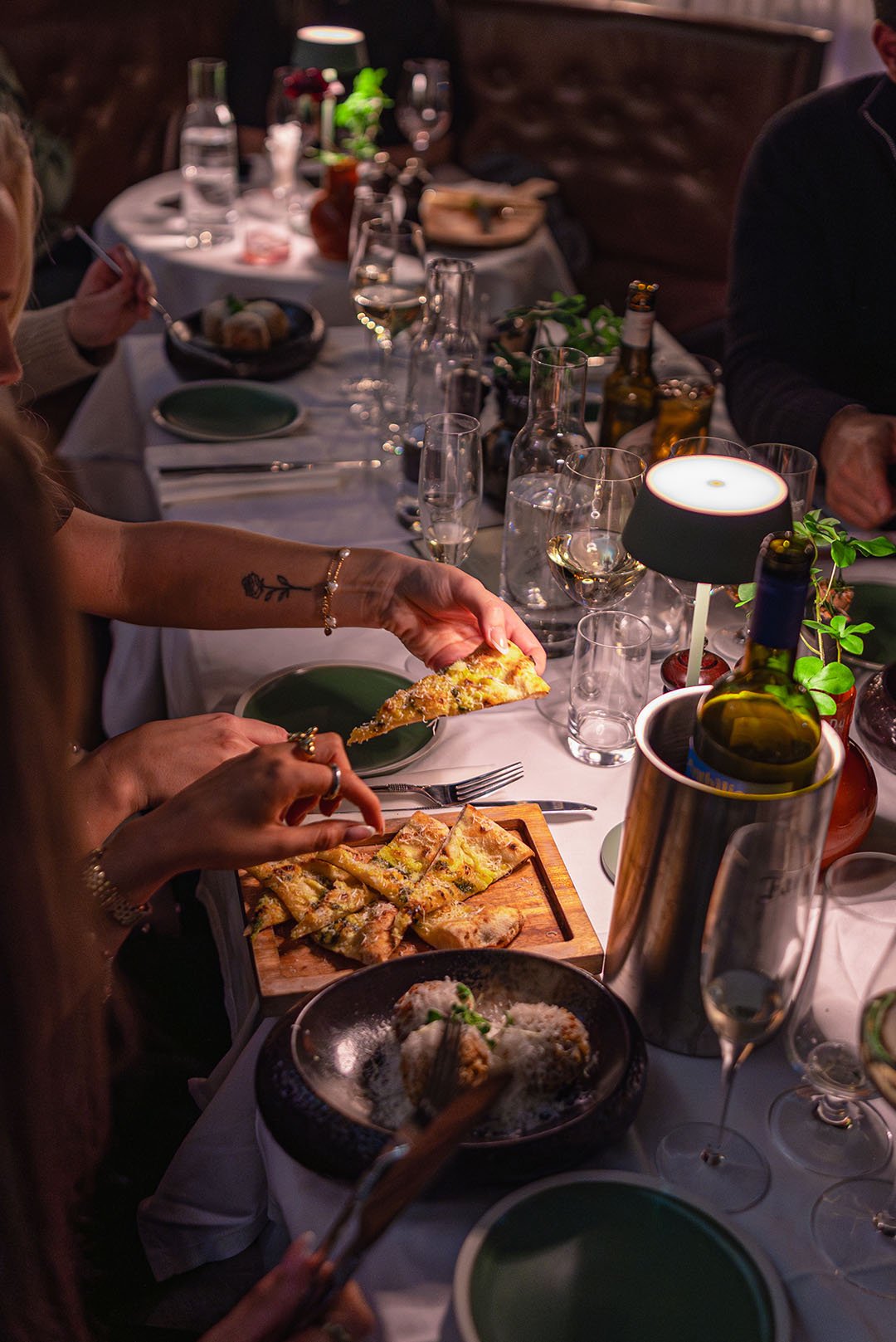 A dinner party with people sharing a variety of dishes and drinks on the table, including pizza, pasta, and bottles of wine. The setting is cozy with warm lighting.