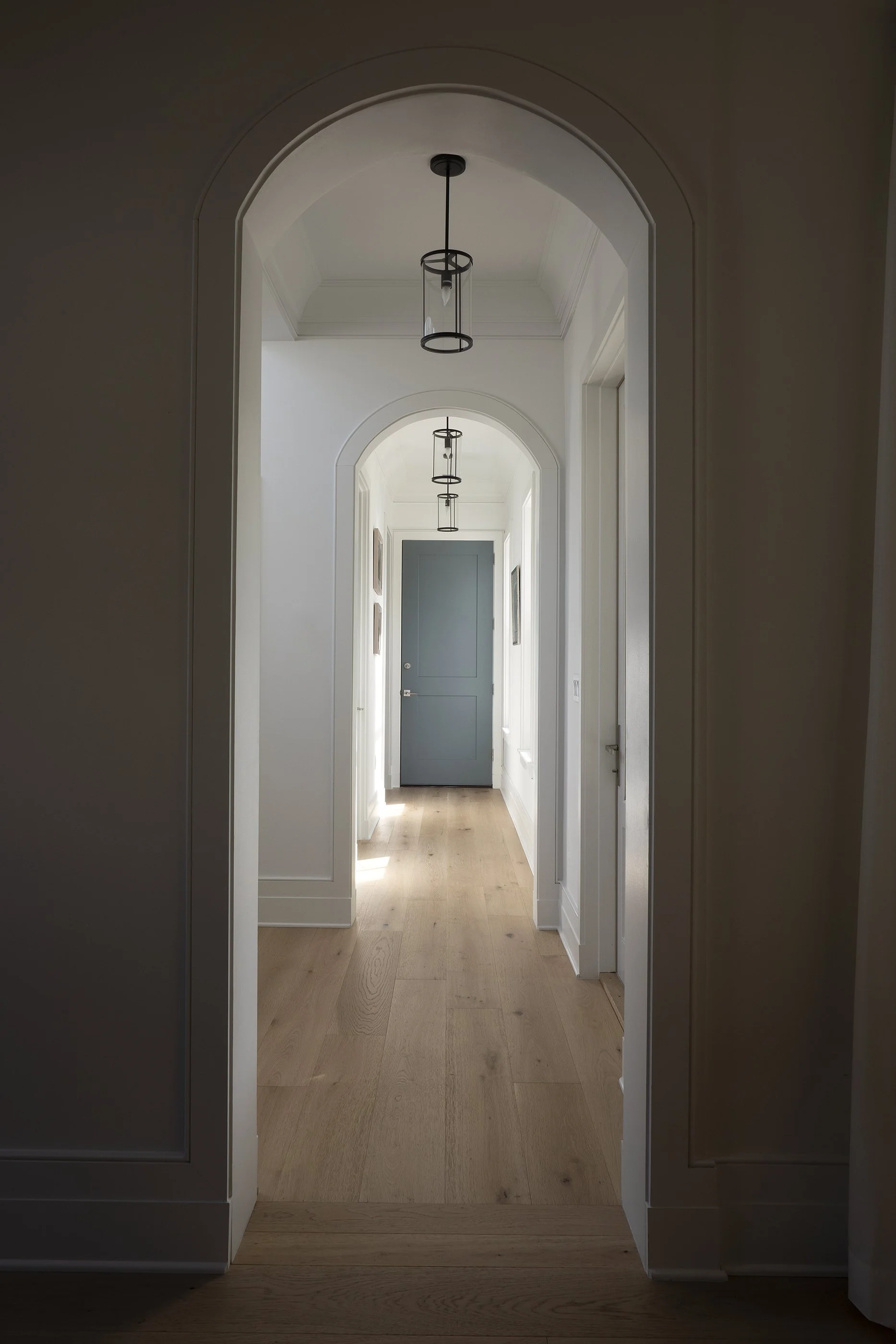 Light-filled hallway with wooden flooring, white walls, and black pendant lights hanging from the ceiling, leading to a blue door at the end.