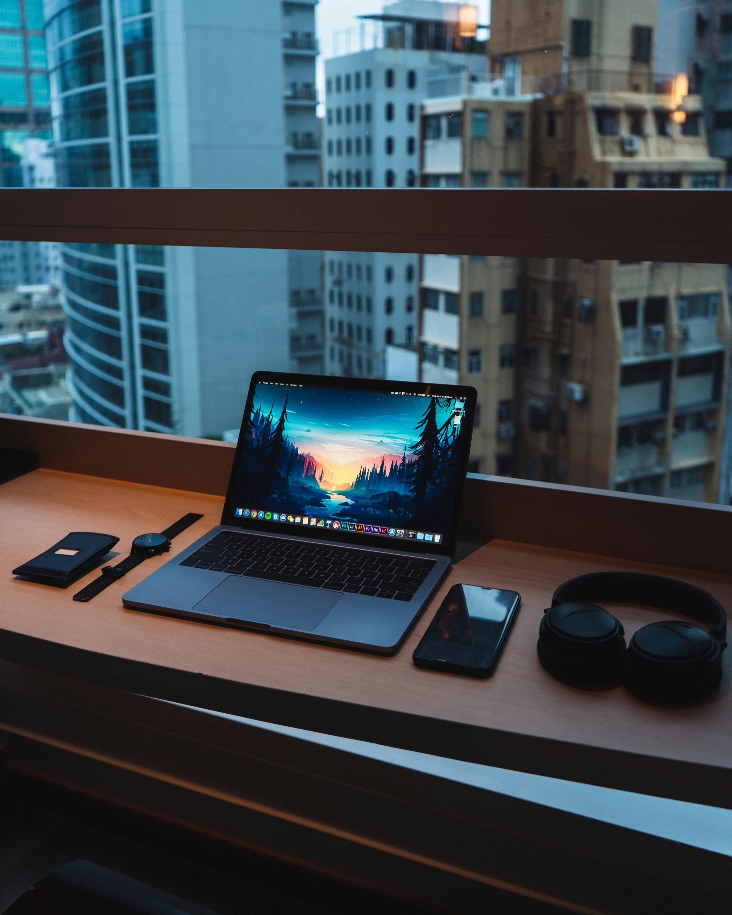 Laptop with landscape wallpaper, smartphone, headphones, watch, and wallet on a desk in a high-rise apartment with city buildings outside window.