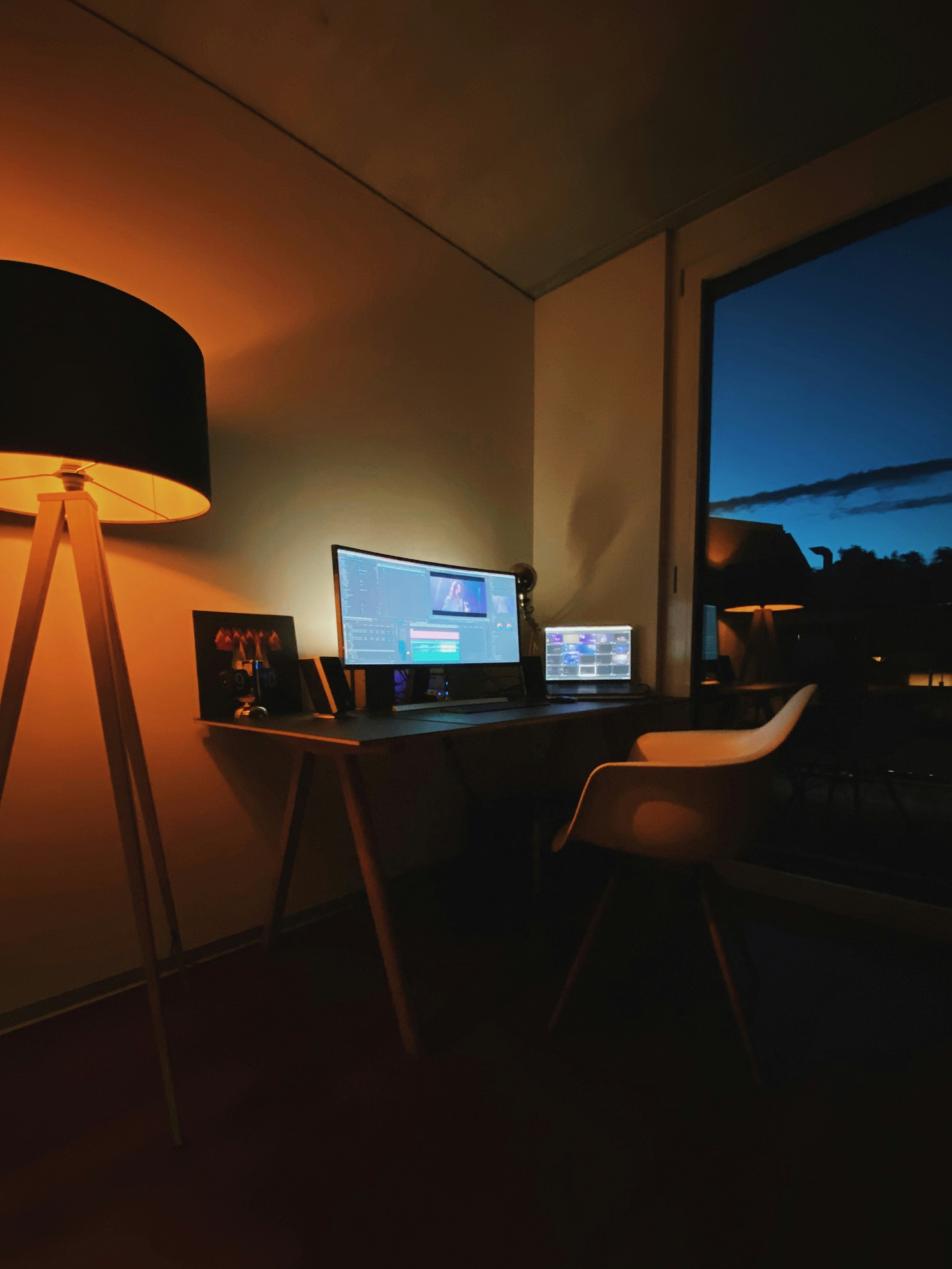 A home office setup at night with a desk, dual monitors, a chair, and a tall floor lamp casting warm light, with a window showing a dark blue evening sky.