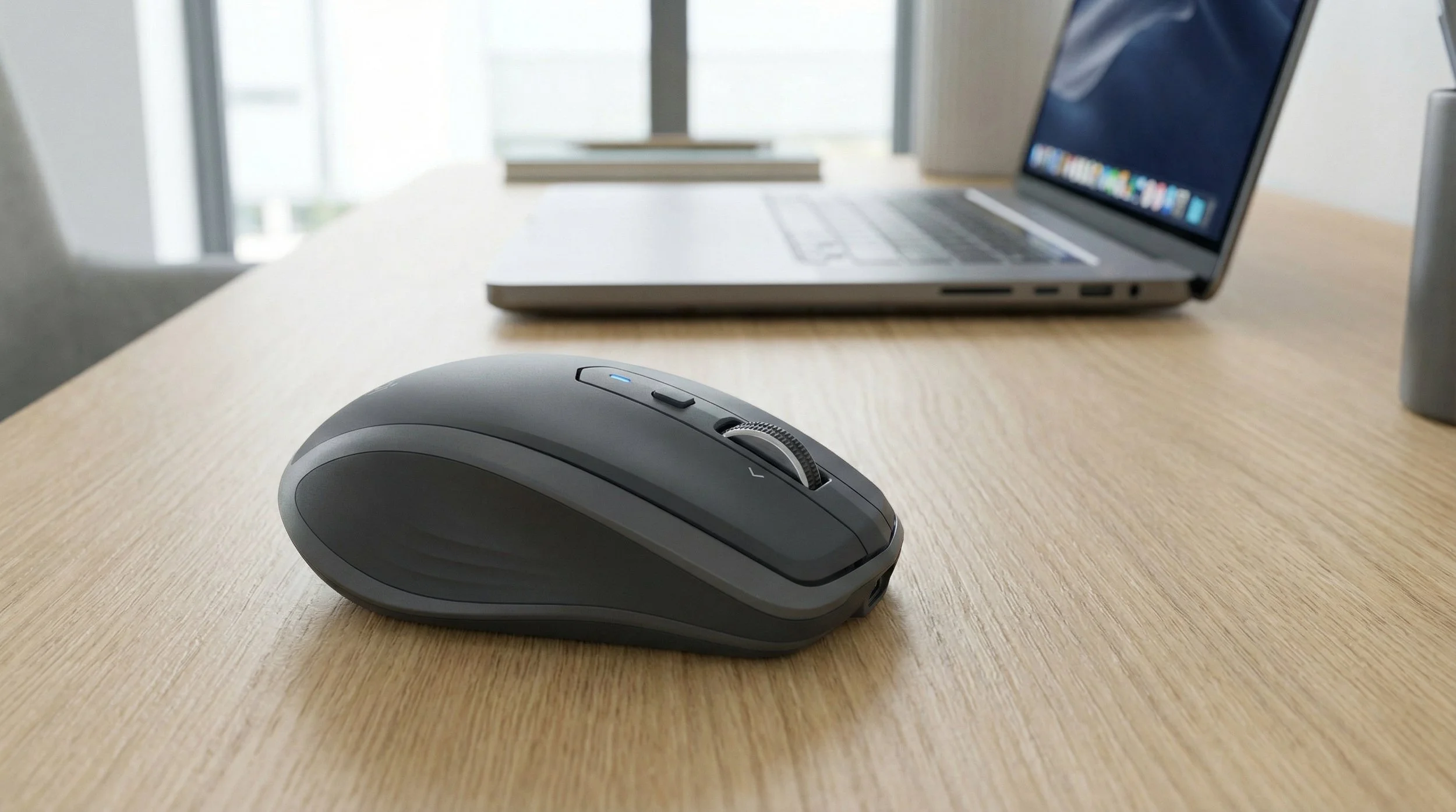 Wireless computer mouse on a wooden desk with an open laptop in the background.