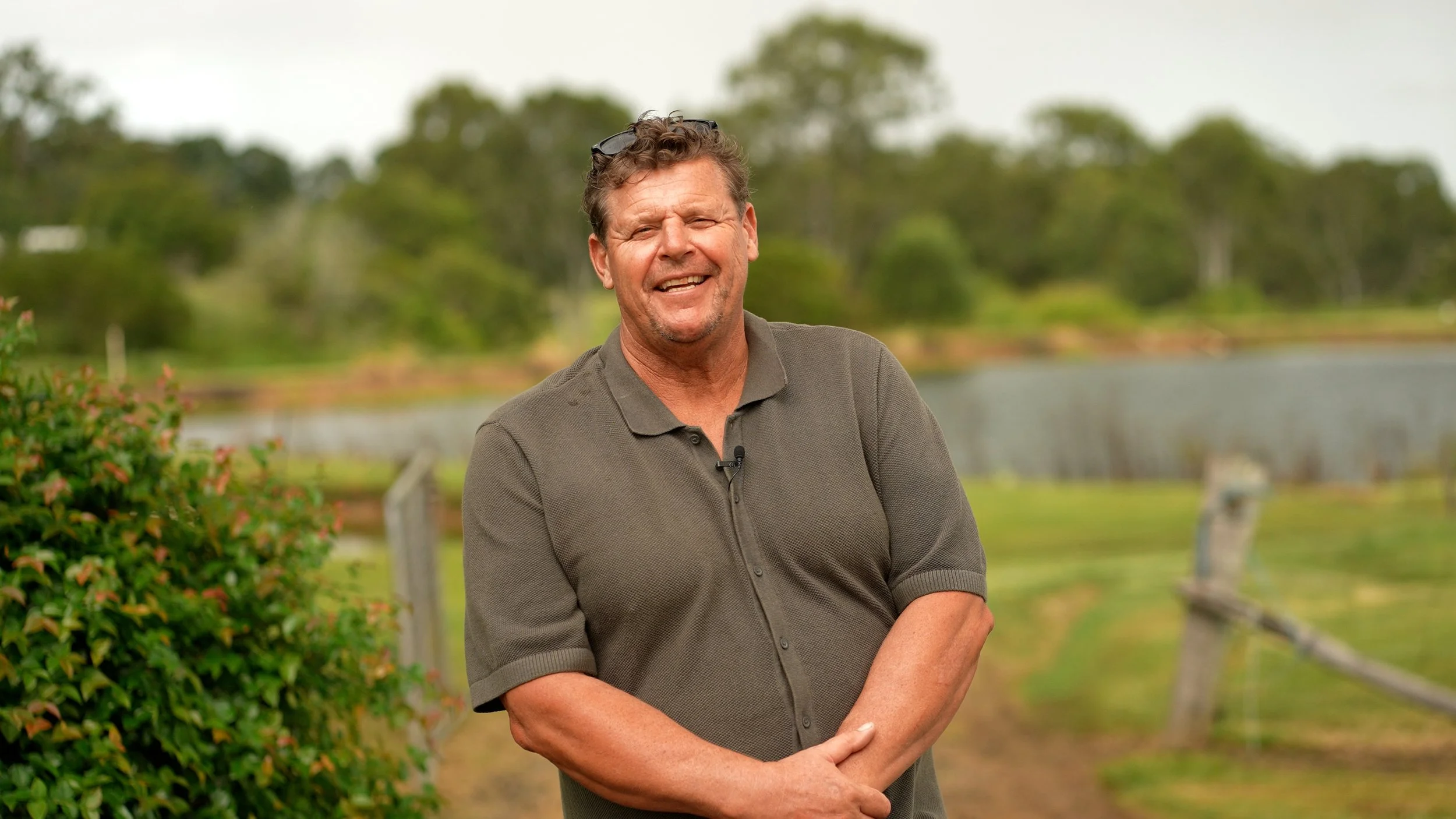 Smiling man with sunglasses on his head, wearing a dark polo shirt, standing outdoors near a body of water with greenery and trees in the background. Fish for Dams.