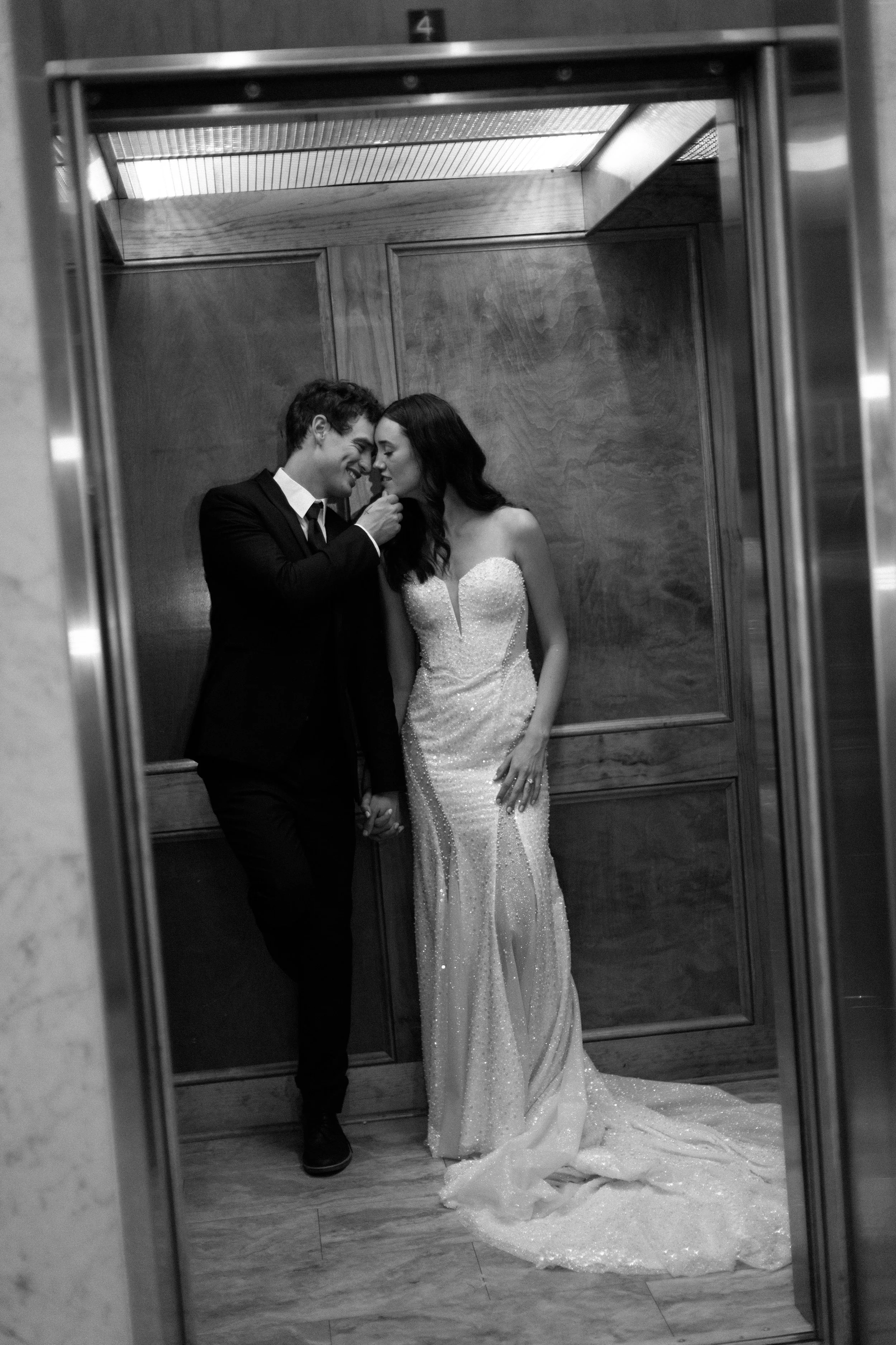 A couple dressed in wedding attire stands kissing in an elevator.