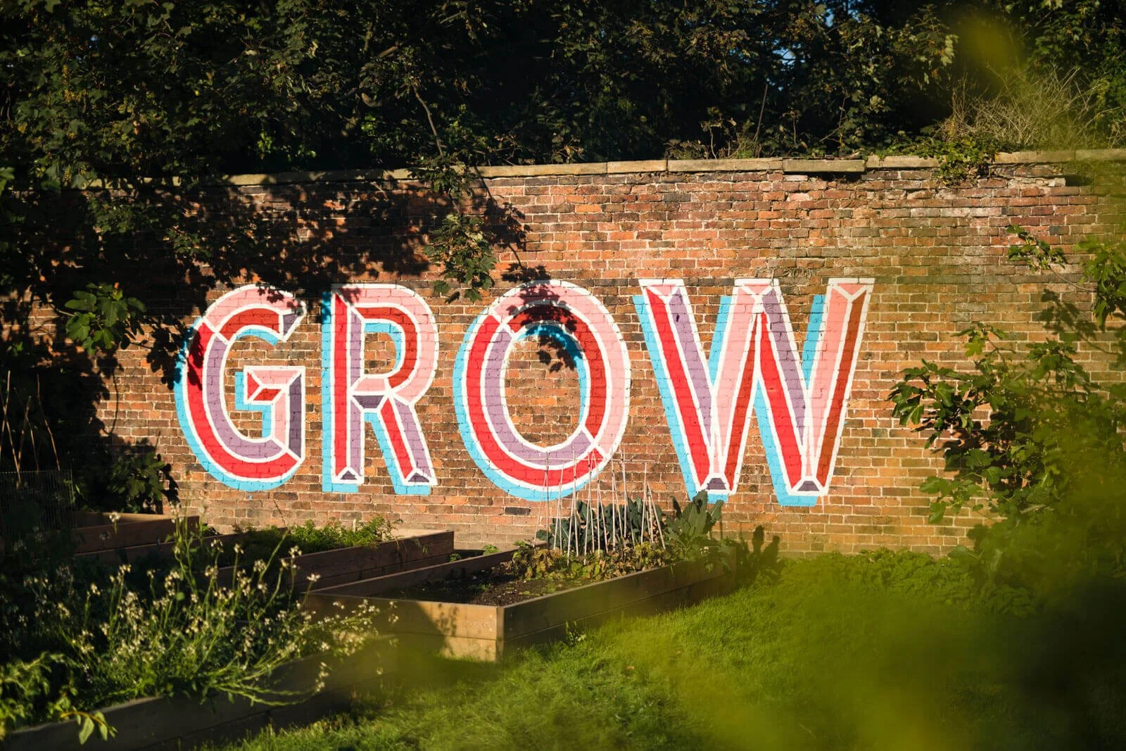 Brick wall with "GROW" painted in colorful letters, surrounded by greenery and garden beds.