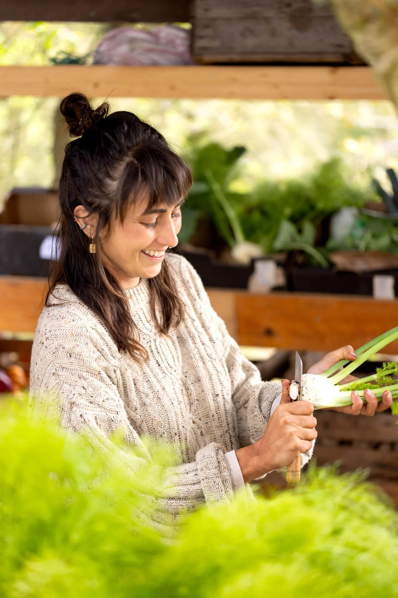 Person peeling a fennel bulb, surrounded by green foliage, wearing a knitted sweater.