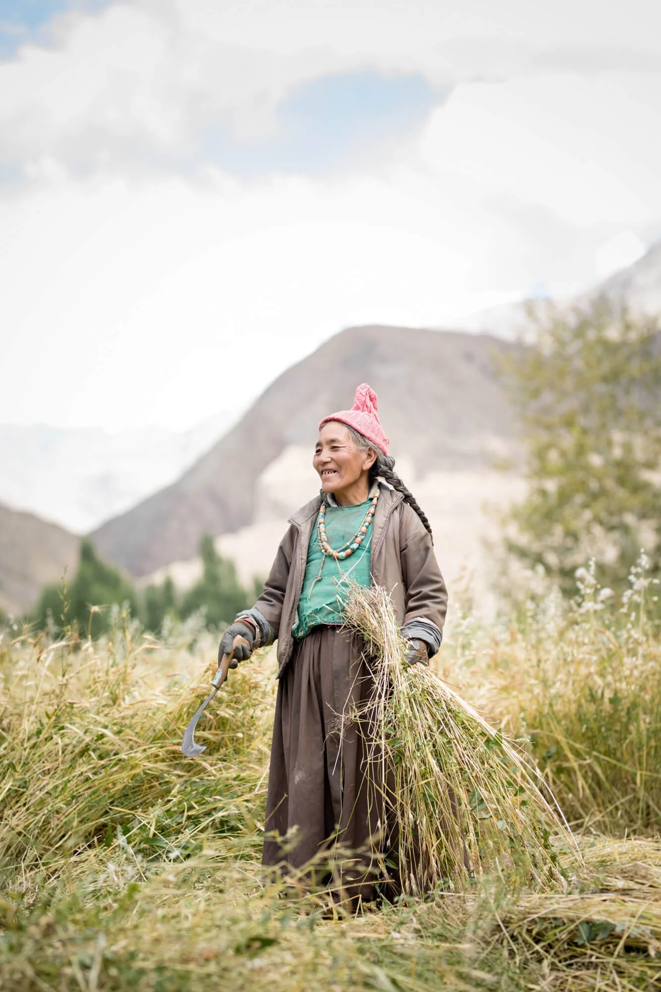 Elderly woman in a field with a sickle and harvested crops, wearing traditional clothing in a rural landscape.
