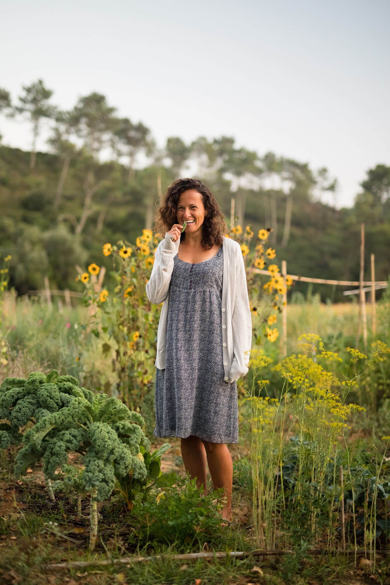 Woman standing in a garden, smiling and holding a vegetable, surrounded by sunflowers and greenery.