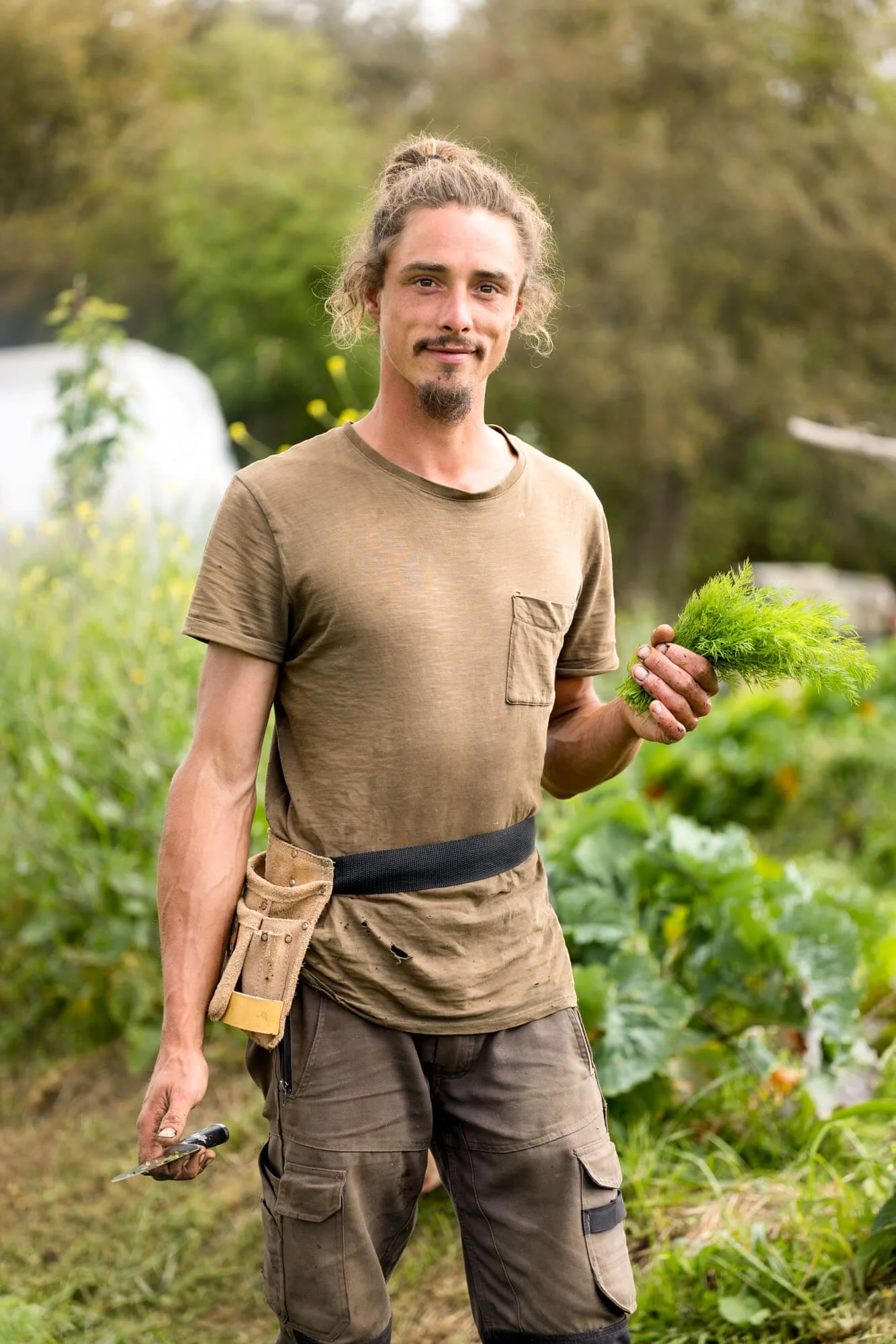 Man with a beard standing in a garden holding plants, wearing a tool belt.