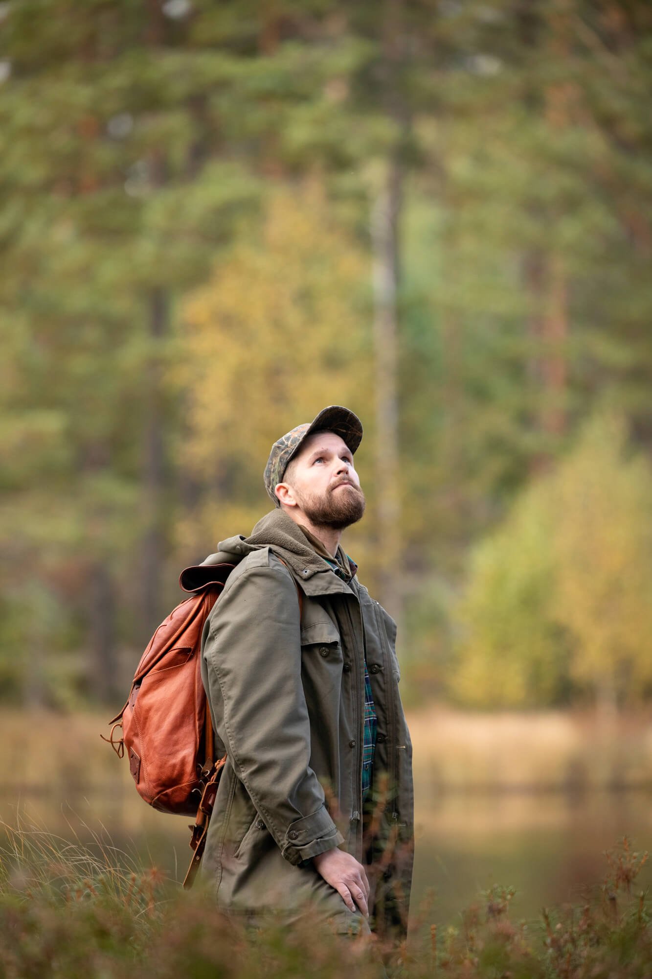 Man in cap and jacket with a backpack standing in a forest, looking upwards.