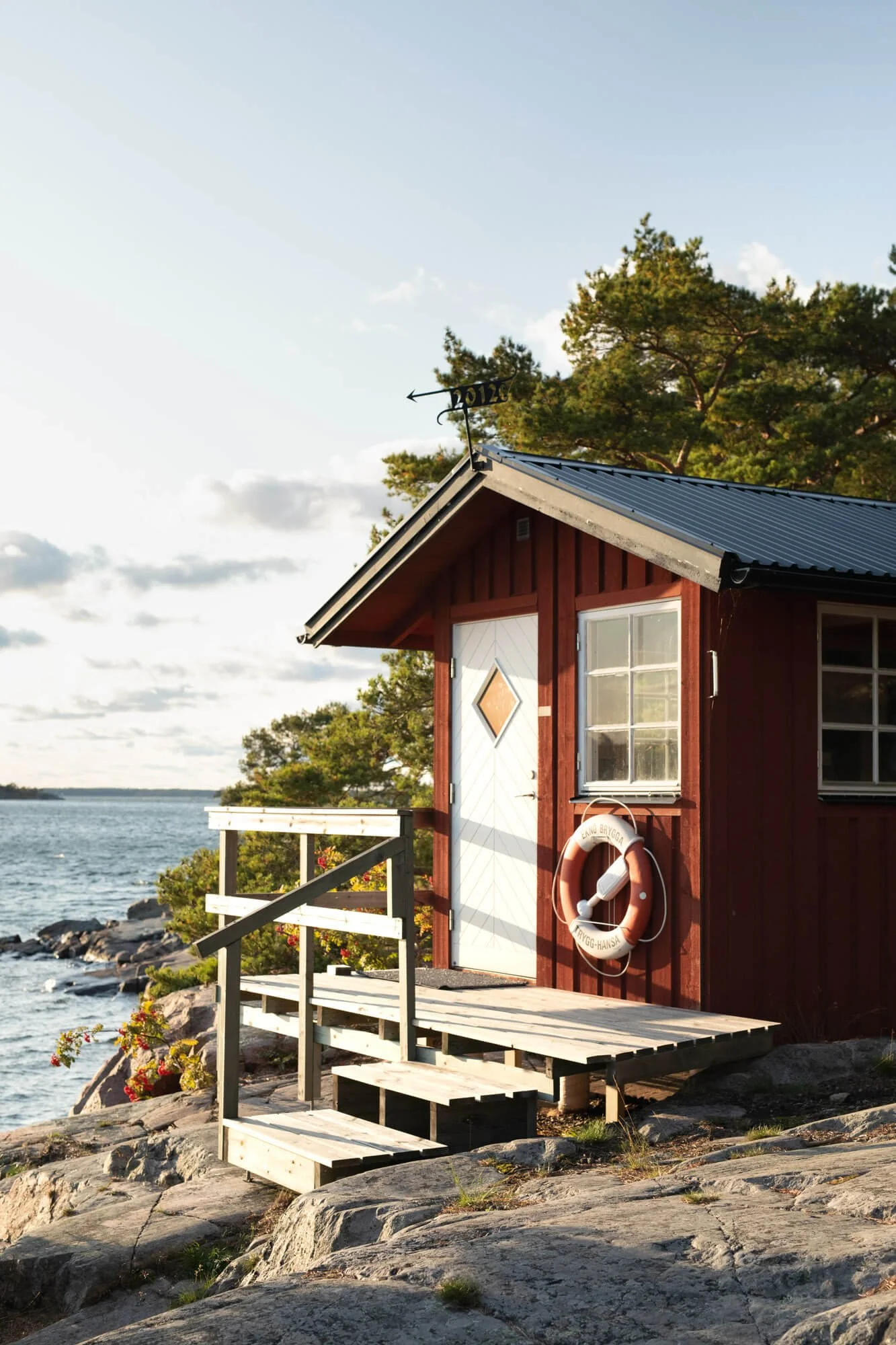 Small red cabin by a rocky coastline with a life preserver attached, surrounded by trees and overlooking the water.