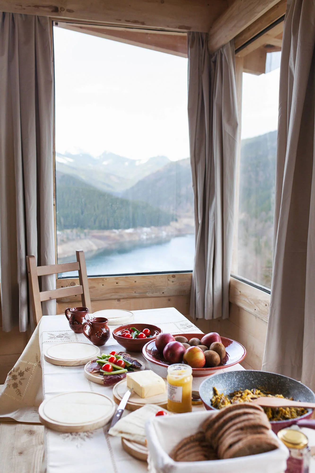 Breakfast table with fruits, bread, cheese, and a view of mountains and a lake through large windows.