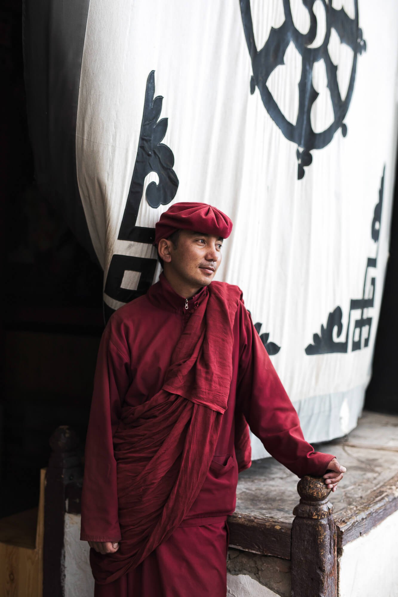 A monk in traditional red attire with a cap standing in front of a large white curtain with black designs.