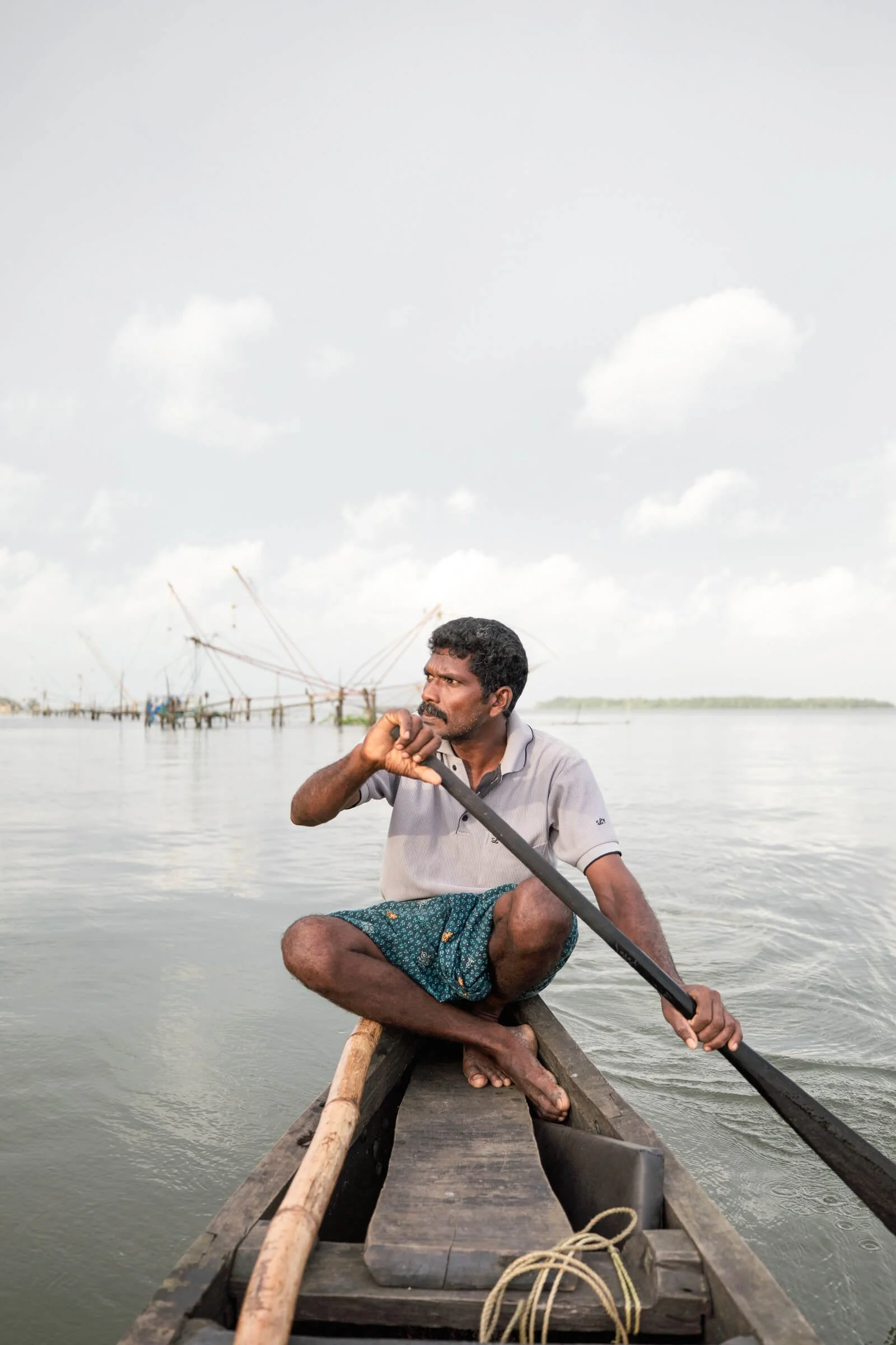 Indian man paddling a wooden boat on water, with fishing nets in the background.