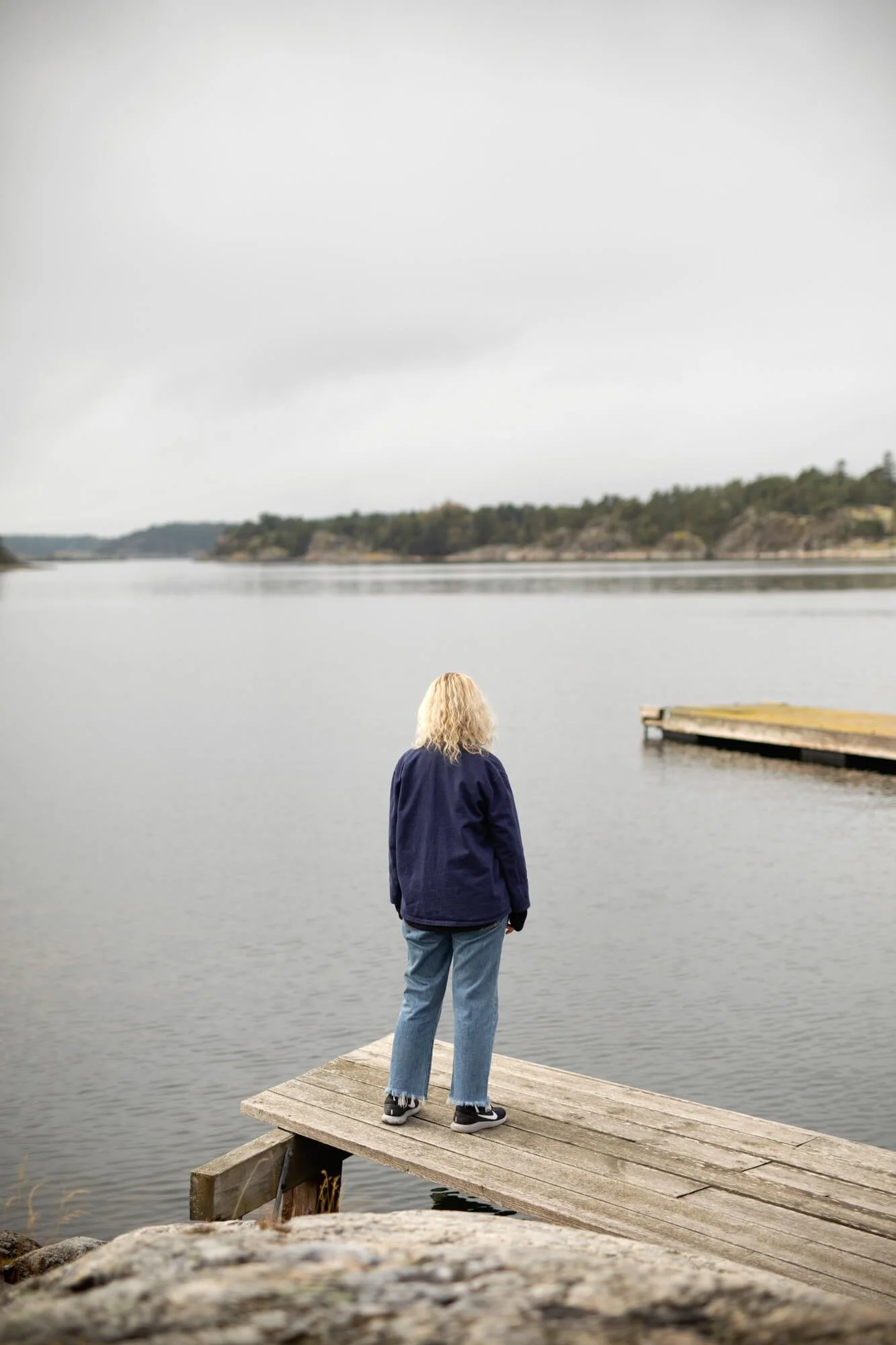 Person with blonde hair standing on a wooden dock overlooking a calm lake with forested hills in the background.