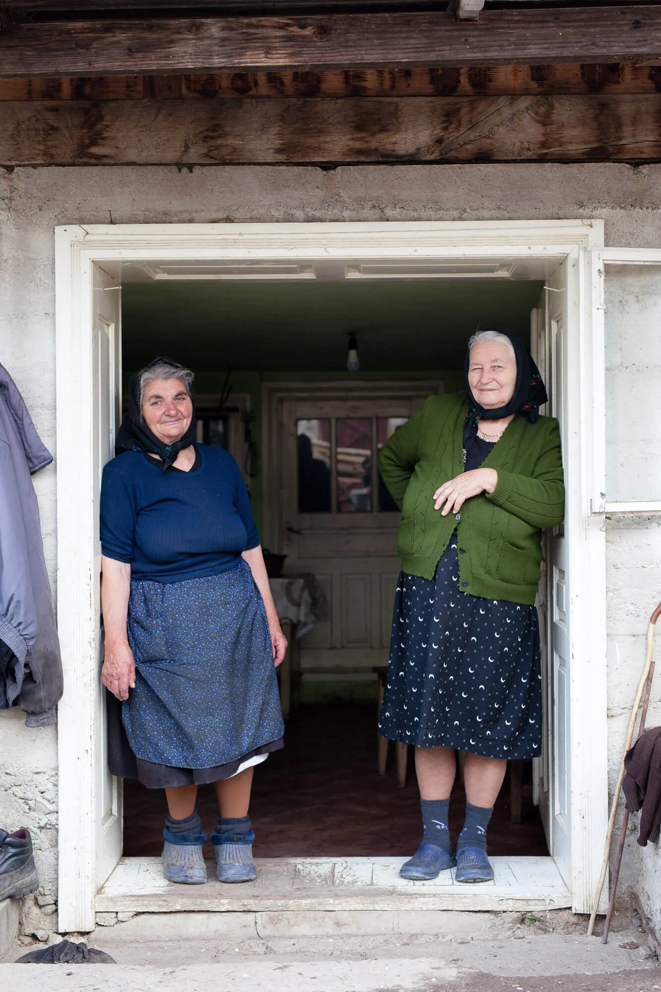 Two elderly women standing in a doorway, wearing headscarves and traditional clothing, smiling at the camera.