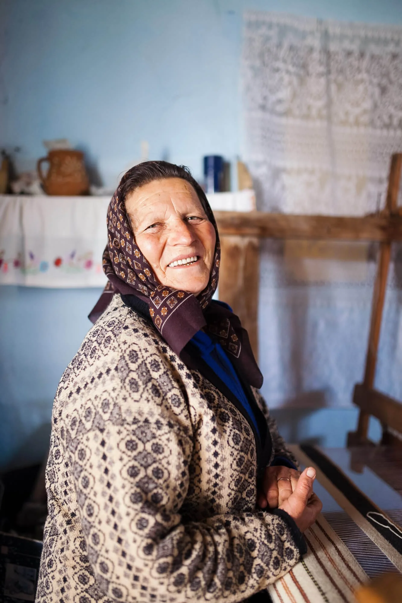 Smiling elderly woman wearing patterned headscarf in cozy room with woven textiles.
