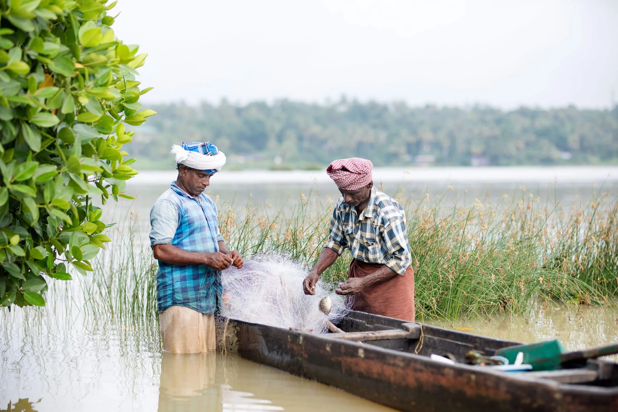 Two men standing in shallow water next to a wooden boat, handling fishing net, surrounded by greenery and reeds.