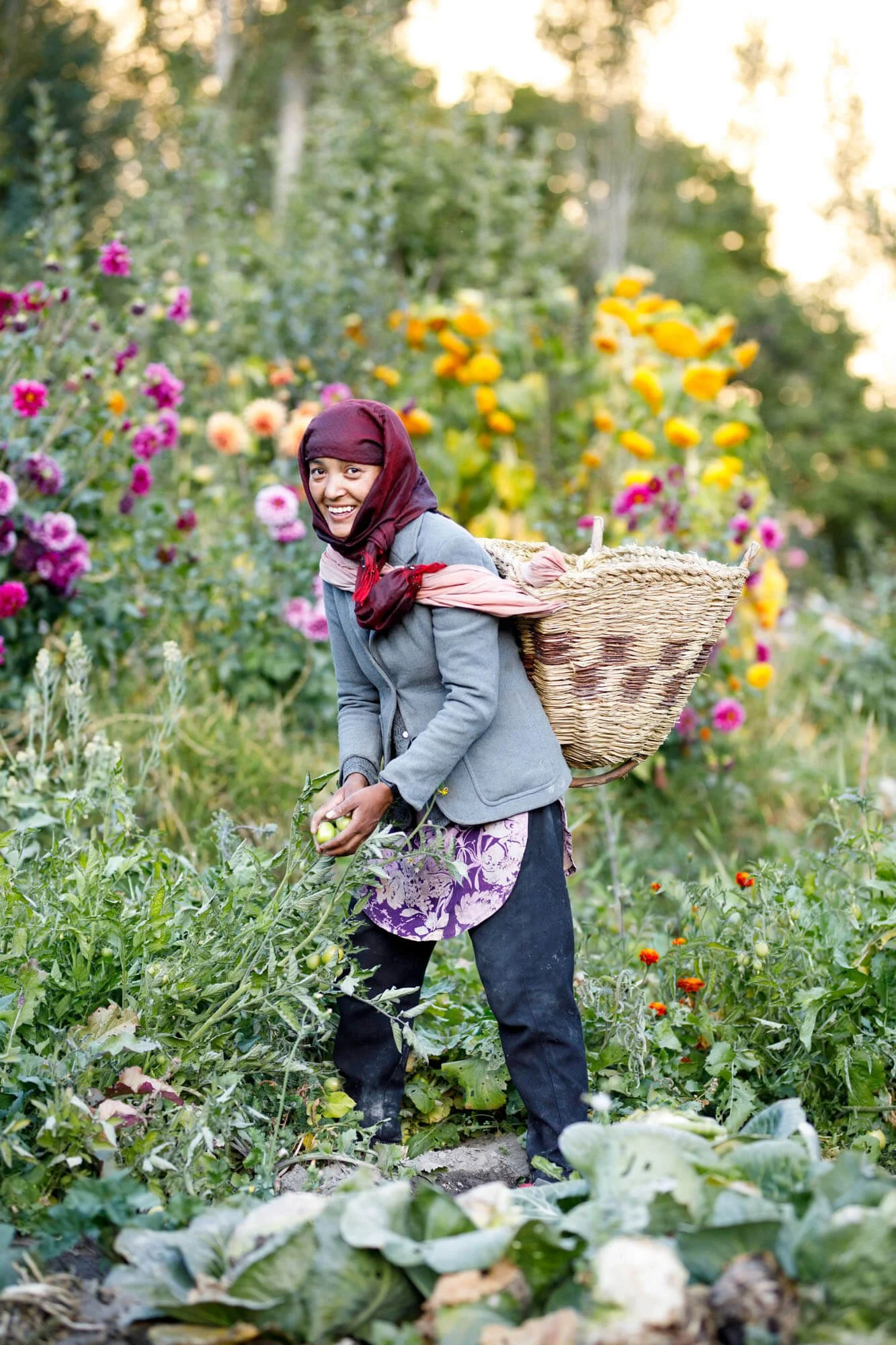 A person in a garden with a headscarf and a woven basket, picking green tomatoes. The background is filled with colorful flowers and lush greenery.