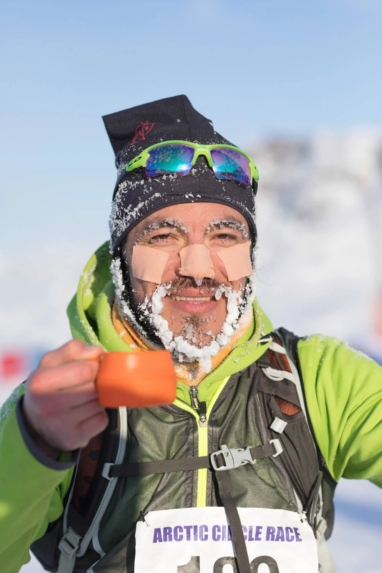 Participant in Arctic Circle Race with frosted beard, wearing green jacket and sunglasses, holding orange cup.