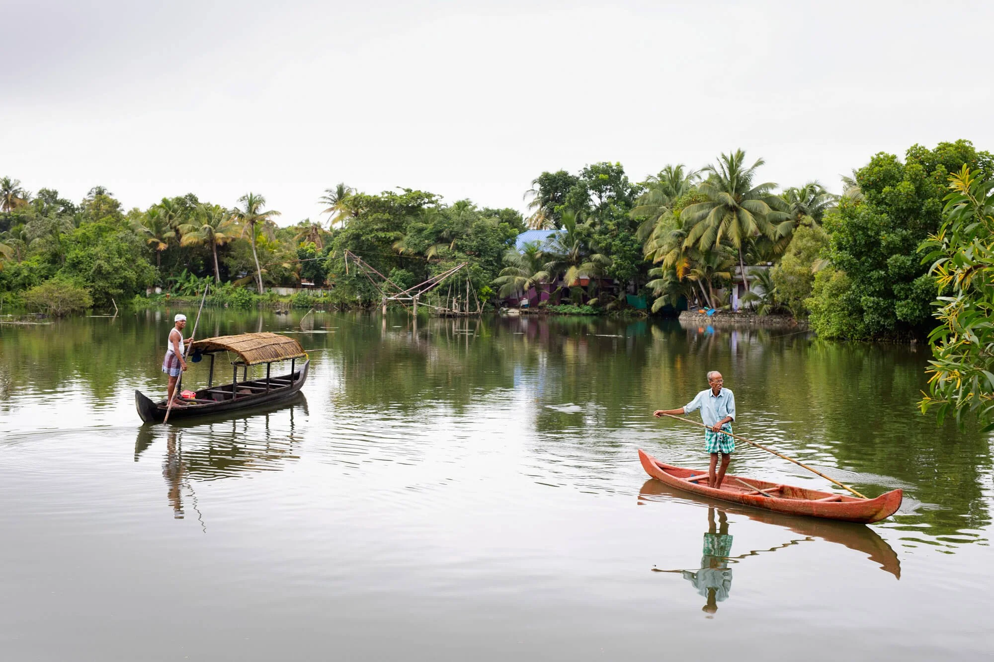 Two men paddling traditional boats on a calm river, surrounded by lush greenery and palm trees.