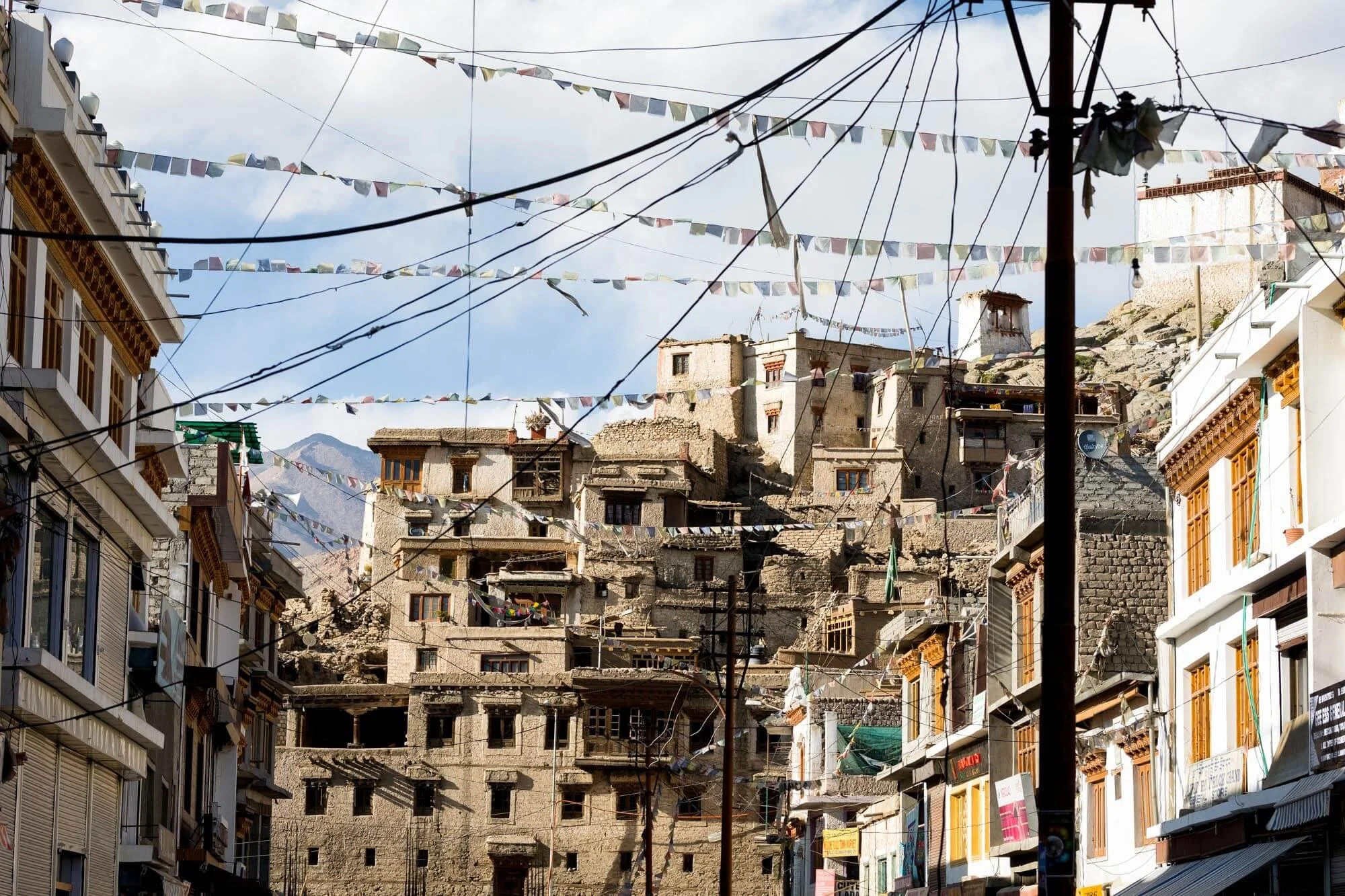 Traditional stone and mud brick buildings in a mountainous area, with prayer flags and electrical wires hanging across the street, under a partly cloudy sky.