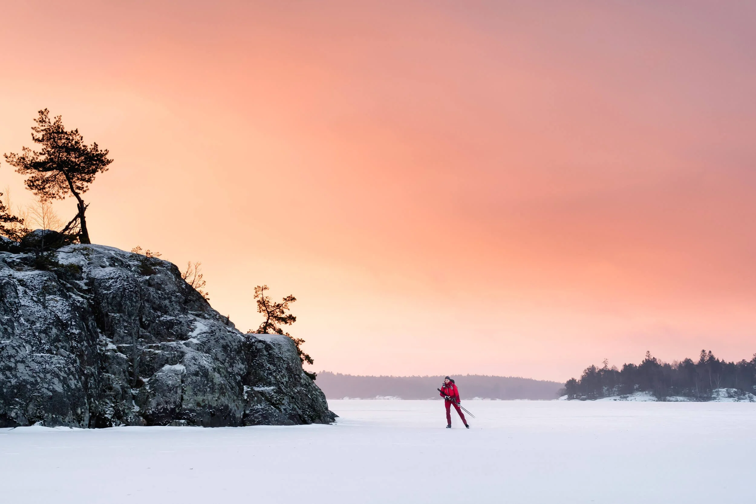 Person in red skiing on a frozen lake with orange sunset sky and rocky terrain.